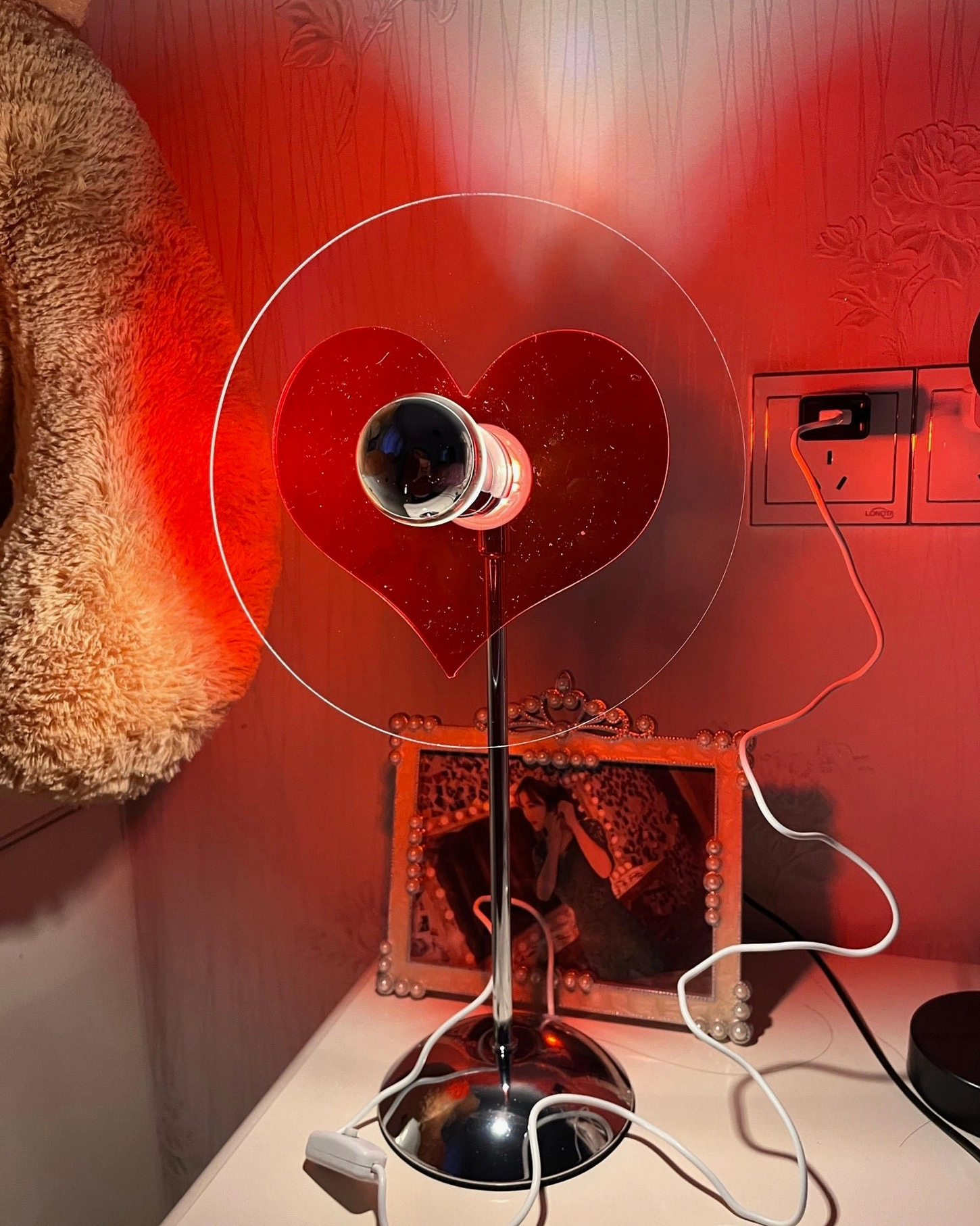 Heart-shaped projection table lamp in bedroom setting, casting warm red heart glow on wall above vanity desk