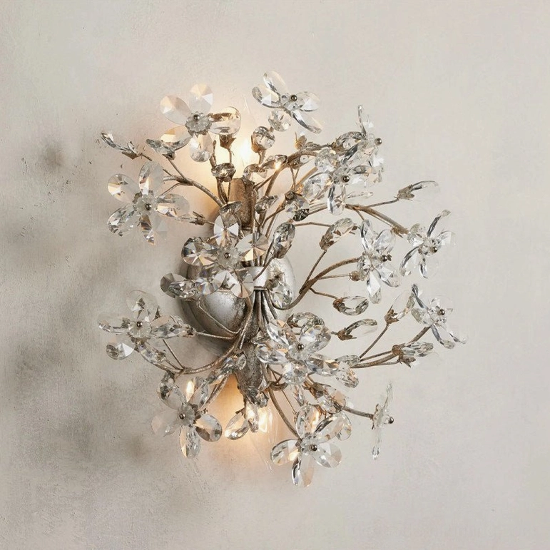 Silver crystal flower wall sconce lit on interior wall, angled view showing clear faceted crystal elements and polished metal branches