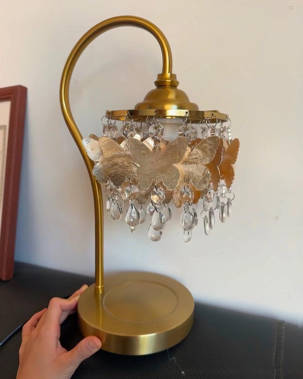 Low side angle view of Jasinski crystal butterfly table lamp next to dark leather sofa, hand touching polished brass base