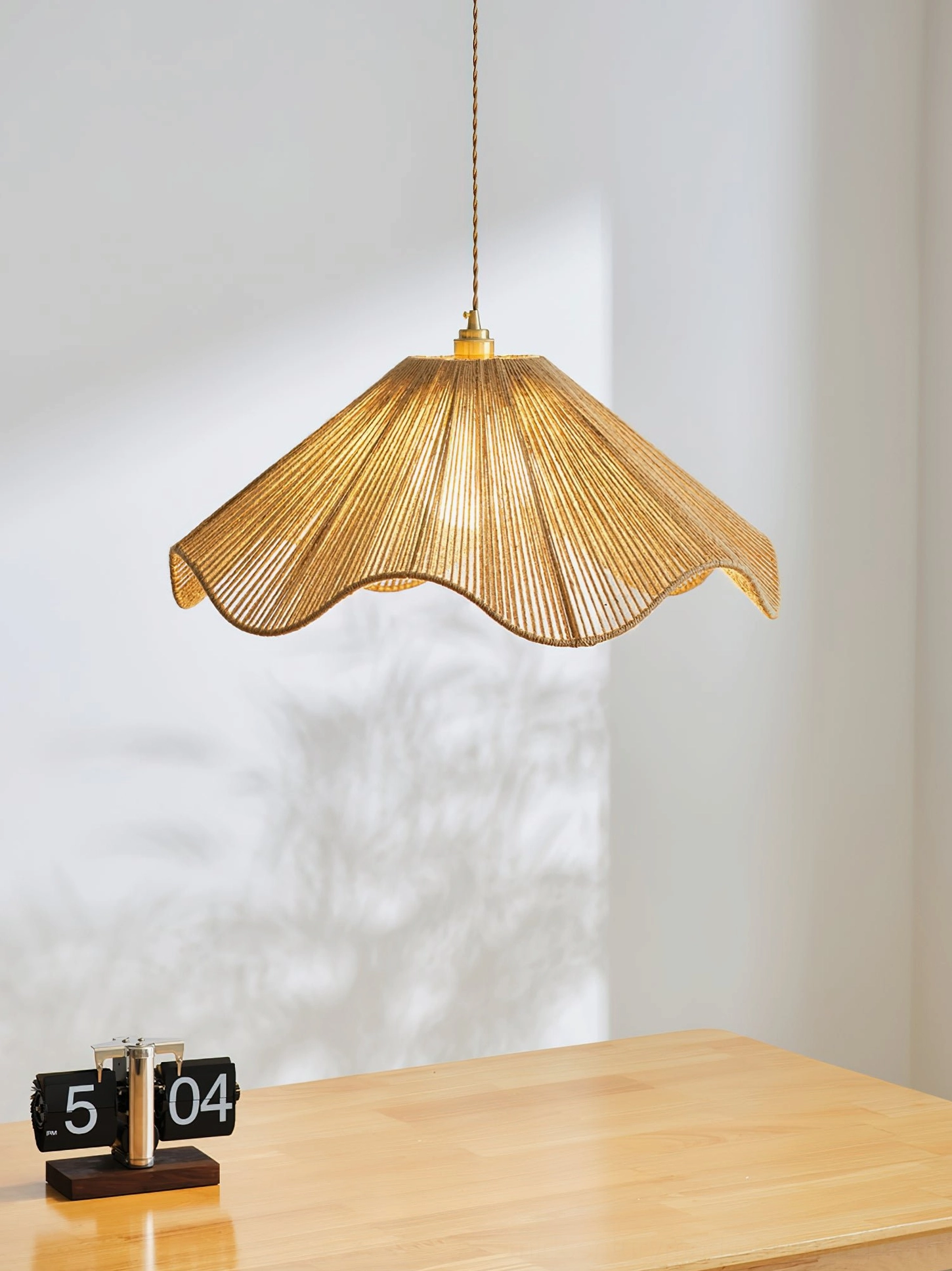 Karai woven pendant light creating soft shadow patterns on wall, installed above minimalist wooden table with clock decor