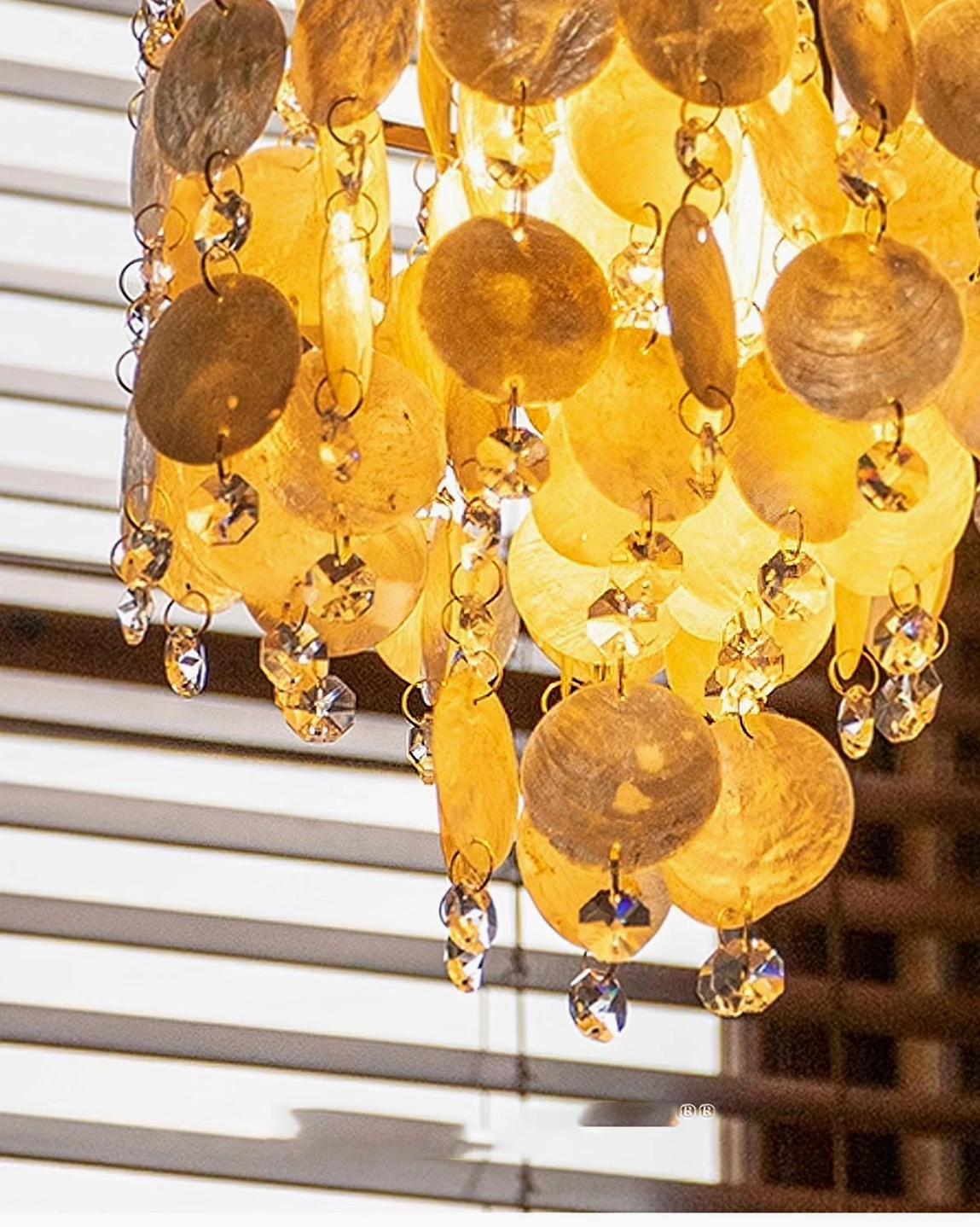 Close-up bottom view of Katinka shell pendant showing layered round shells, tiny crystal charms, warm glowing ambiance near wooden window blinds
