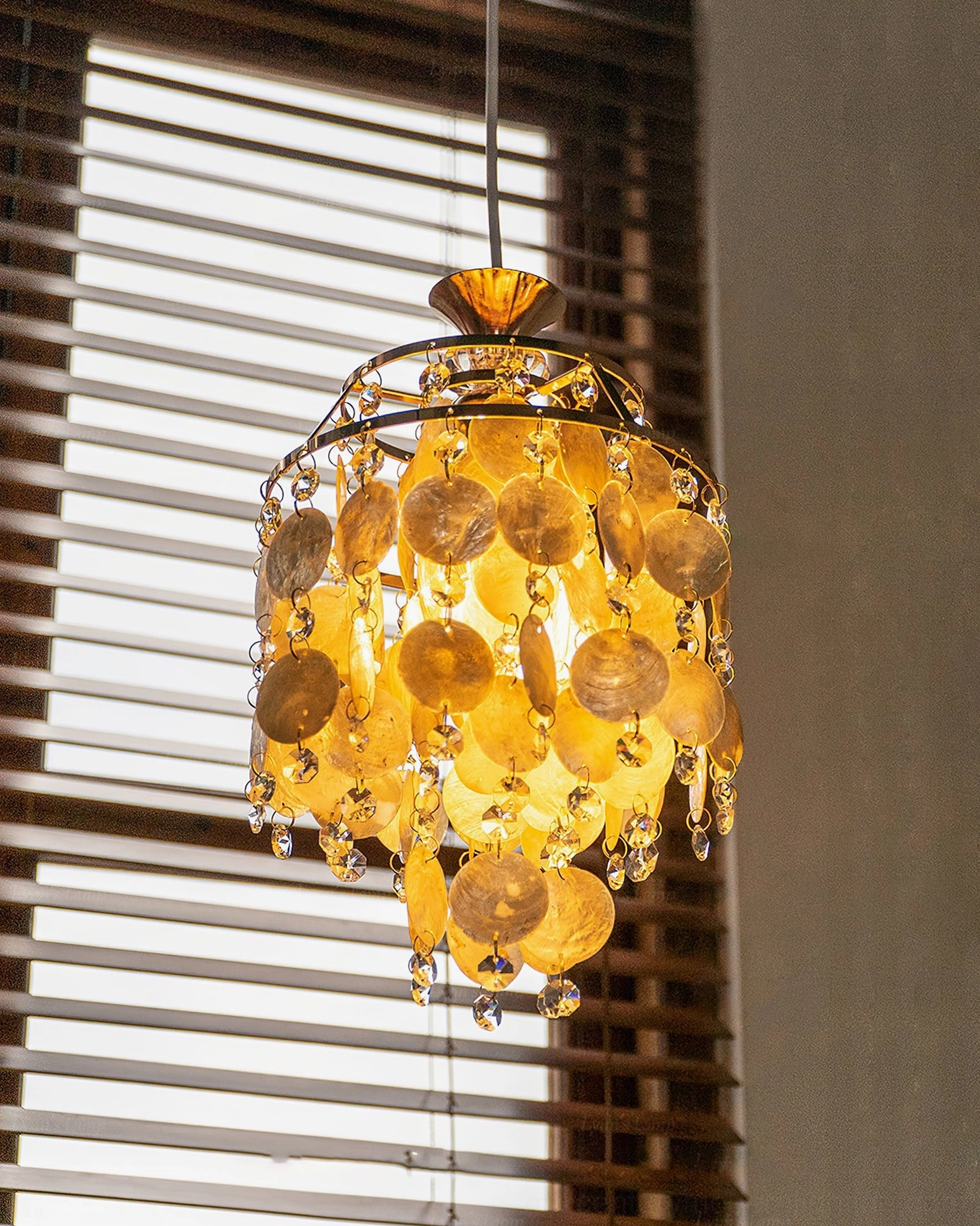 Yellow-brown capiz shell pendant Katinka chandelier fully lit with gold accents, dense layered shells, front side view with louvered window