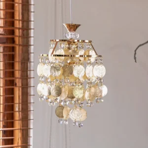 Tiered Katinka pendant chandelier with warm gold finish and mixed clear and beige shells, close-up front view with indoor window blinds background