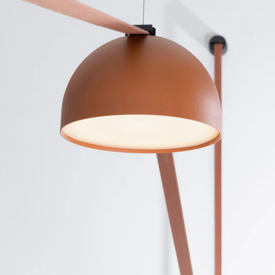 Illuminated matte tan metal dome shade of leather sling pendant lamp glowing against white wall