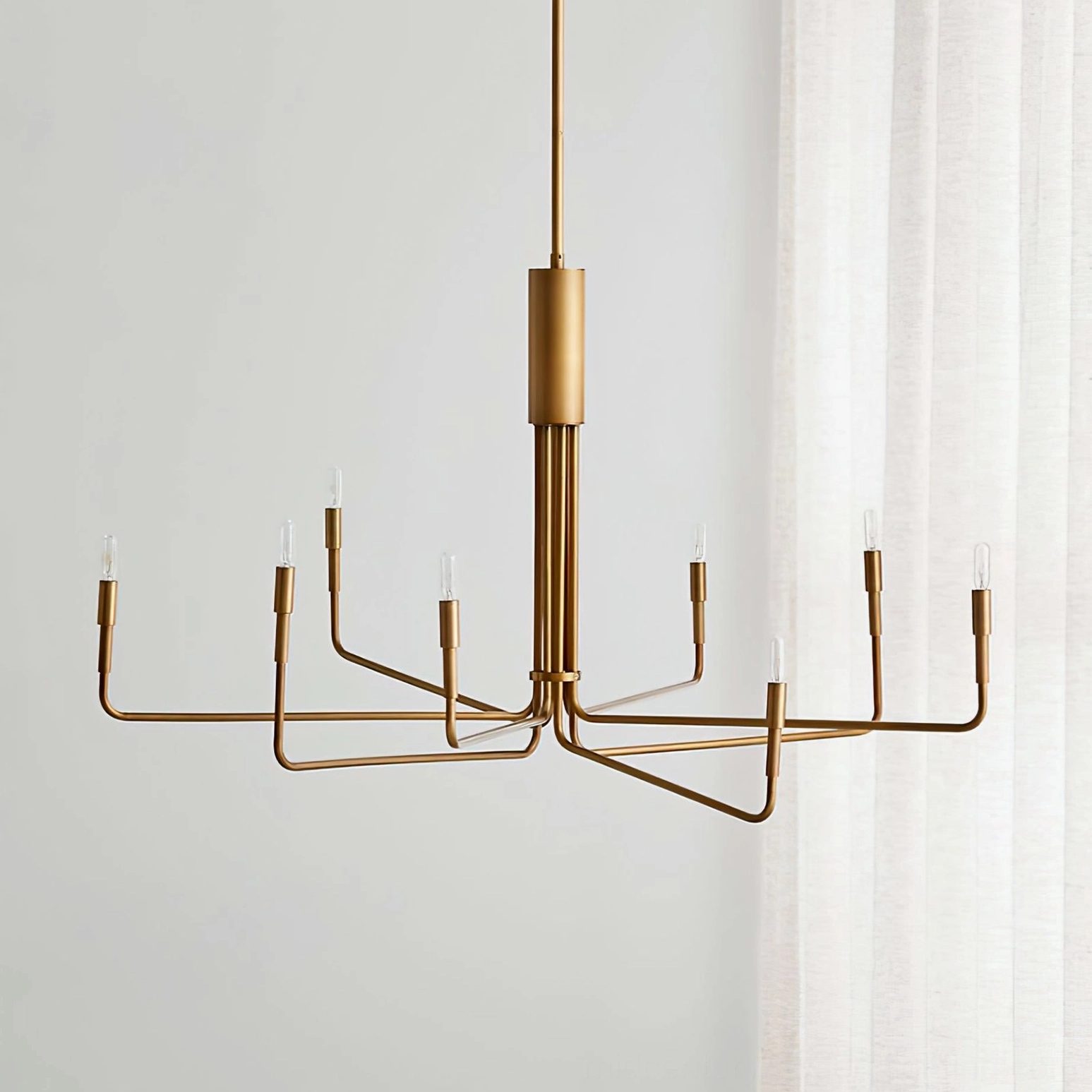 Lepanto brass chandelier angled front view showcasing warm matte brass finish and slim metal arms suspended from ceiling in bright residential dining room with white curtains
