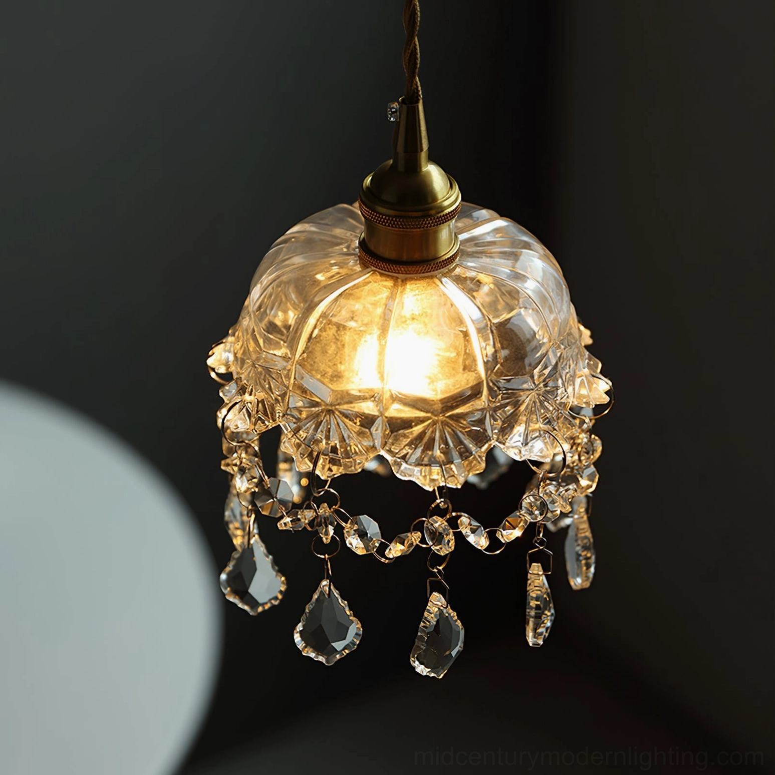 Lit vintage crystal glass pendant with warm bulb glow, brass hardware detail, close-up view
