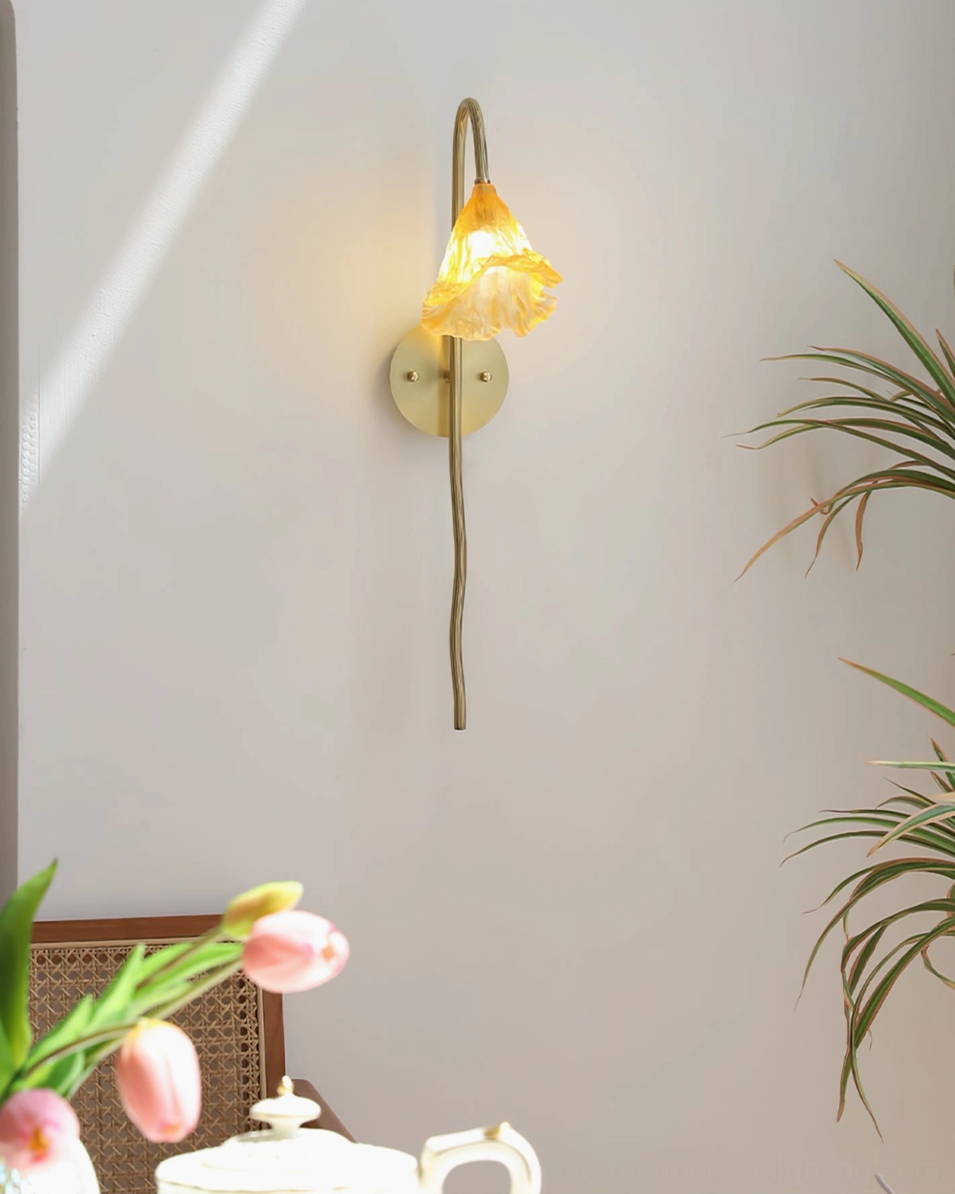 Illuminated Lotus Amber Wall Sconce with warm glowing amber shade on white wall with green plant and tabletop teapot, boho design