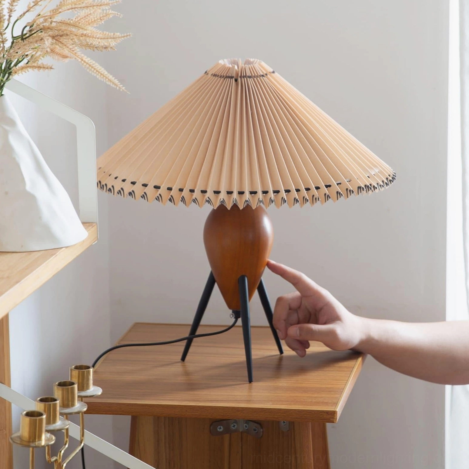 Makie tripod table lamp detail showing hand touching wooden base and metal legs, with dried floral decor