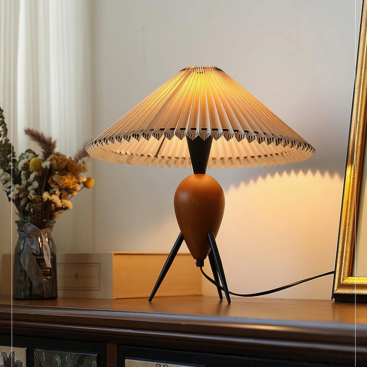 Lit Makie pleated shade tripod lamp on retro dresser creating warm ambient lighting with dried flowers and mirror
