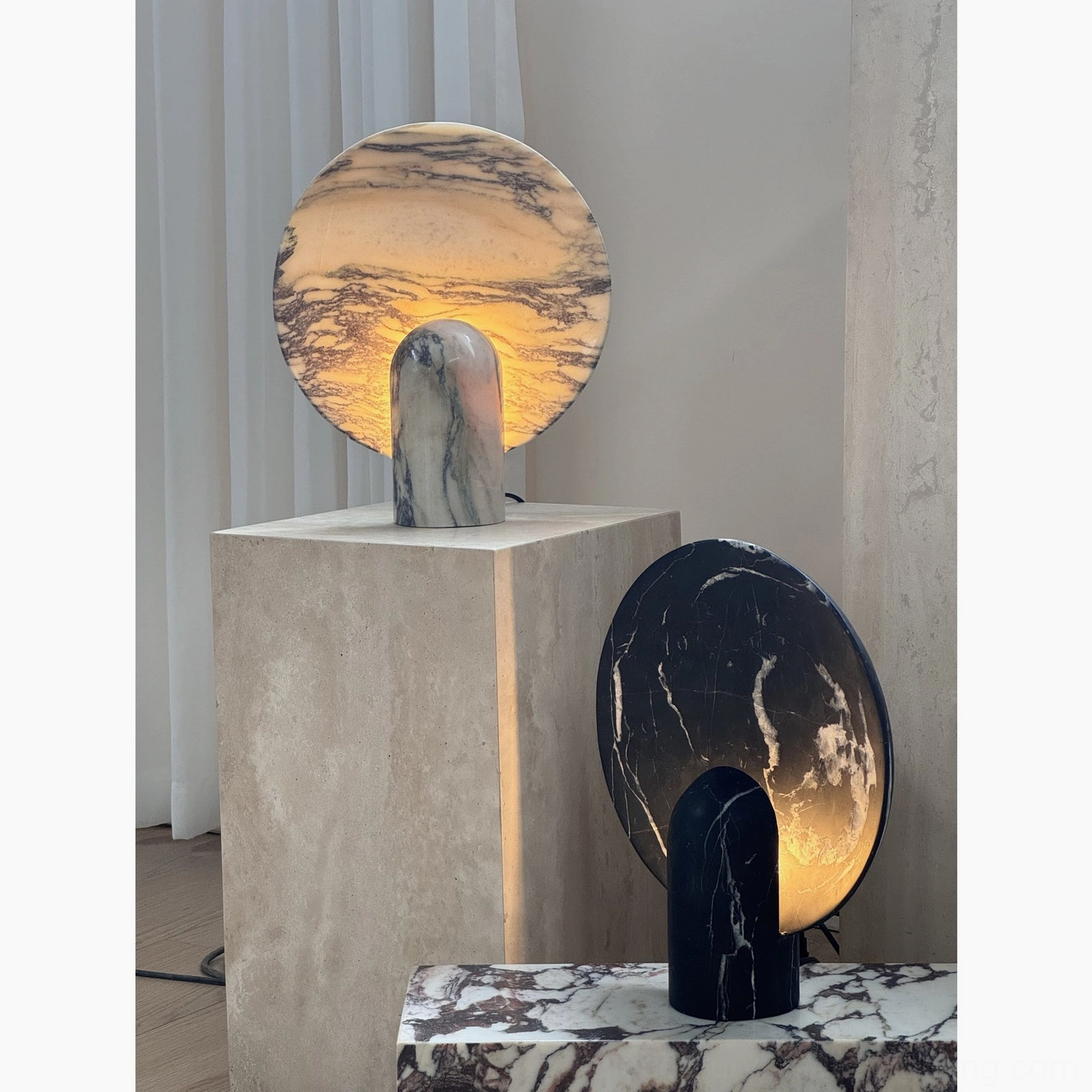 Two marble table lamps with dark veining on concrete studio plinth showing warm glowing illuminated interior light creating ambient elegance
