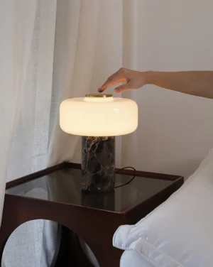 Hand adjusting marble table lamp in bedroom, side eye-level perspective on dark wood bedside table with sheer window curtains