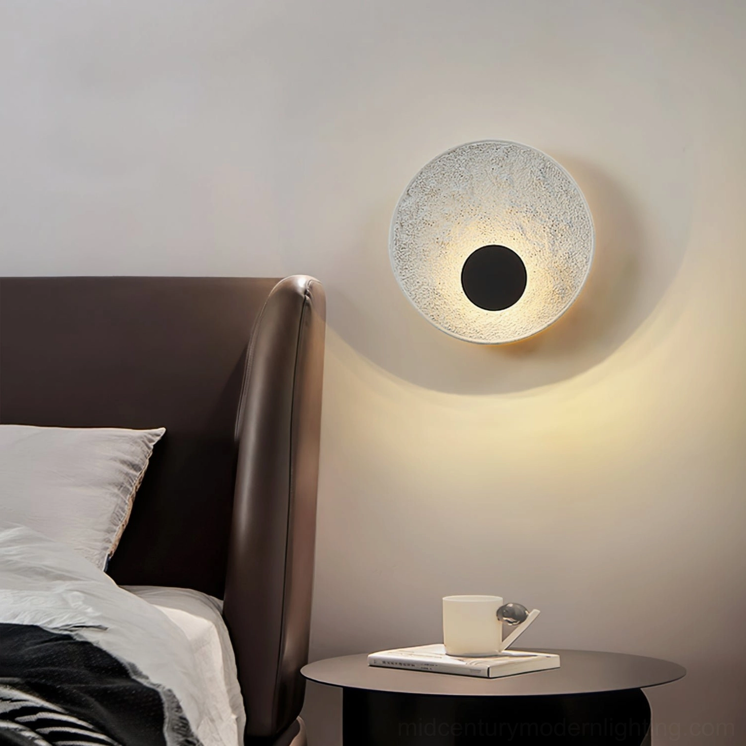 Melesa circular wall sconce installed in bedroom with dark leather headboard and contemporary styling