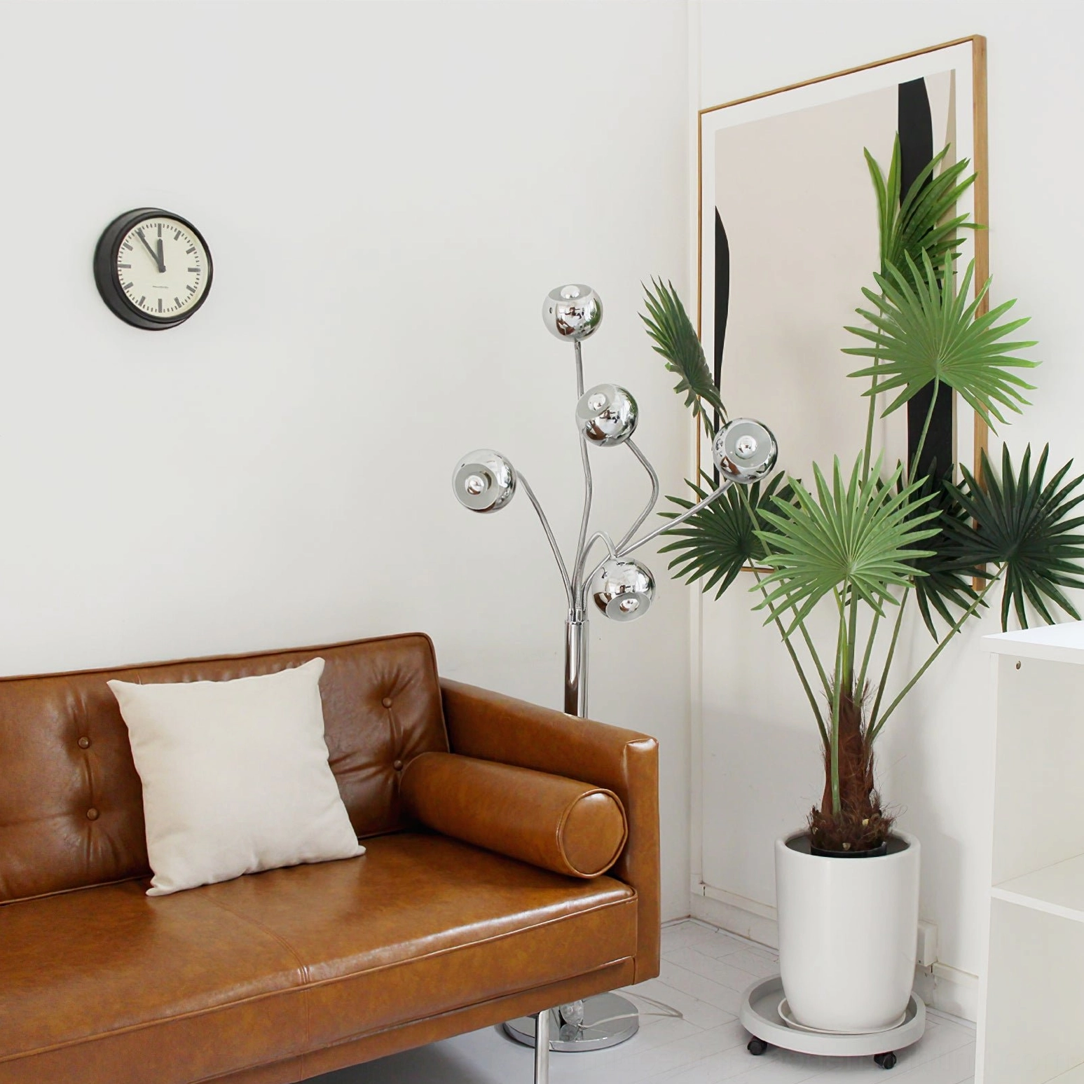 Chrome eyeball floor lamp in mid-century modern living room beside brown leather sofa with potted palm plant and wall clock