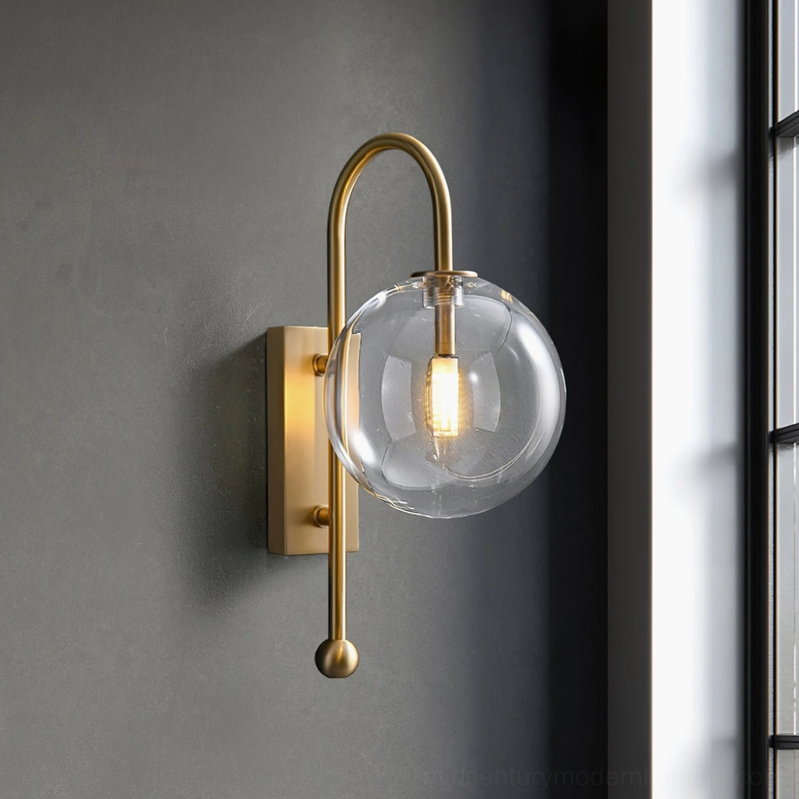 Mid-century modern brass wall sconce with spherical glass shade casting warm light against window