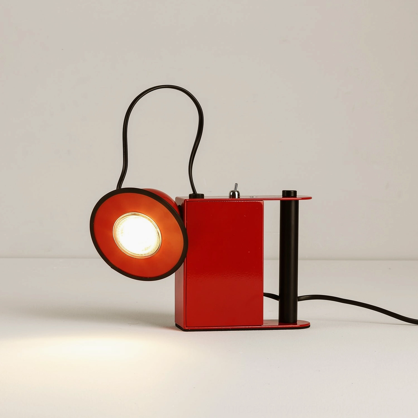 Minibox spotlight table lamp in red colorway, front-facing view with warm illumination and black handle accent
