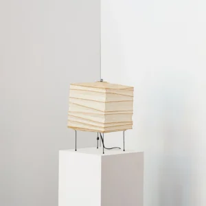 Organic paper table lamp at three-quarter angle on minimalist white display pedestal, showcasing perforated shade detail and ambient light diffusion