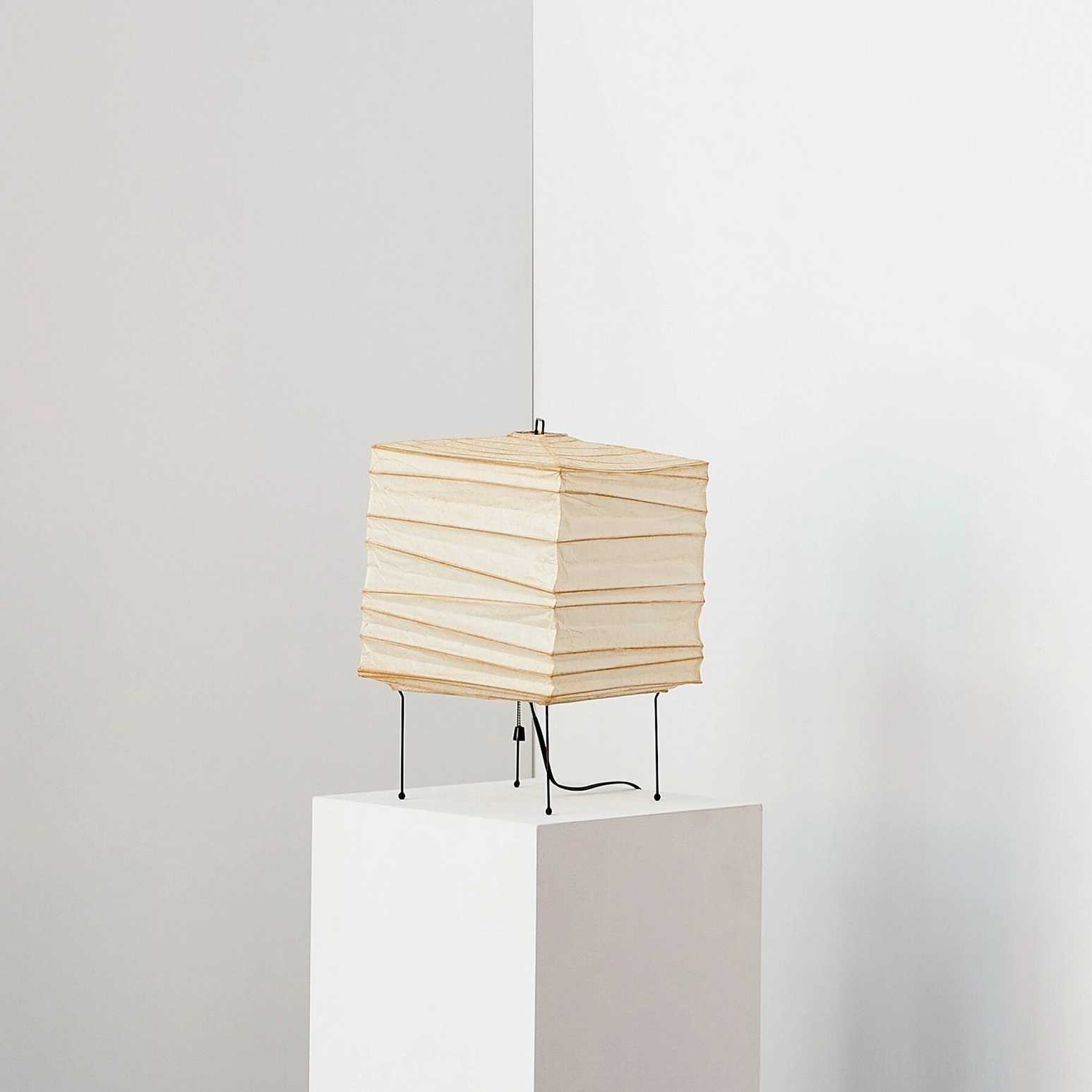 Organic paper table lamp at three-quarter angle on minimalist white display pedestal, showcasing perforated shade detail and ambient light diffusion