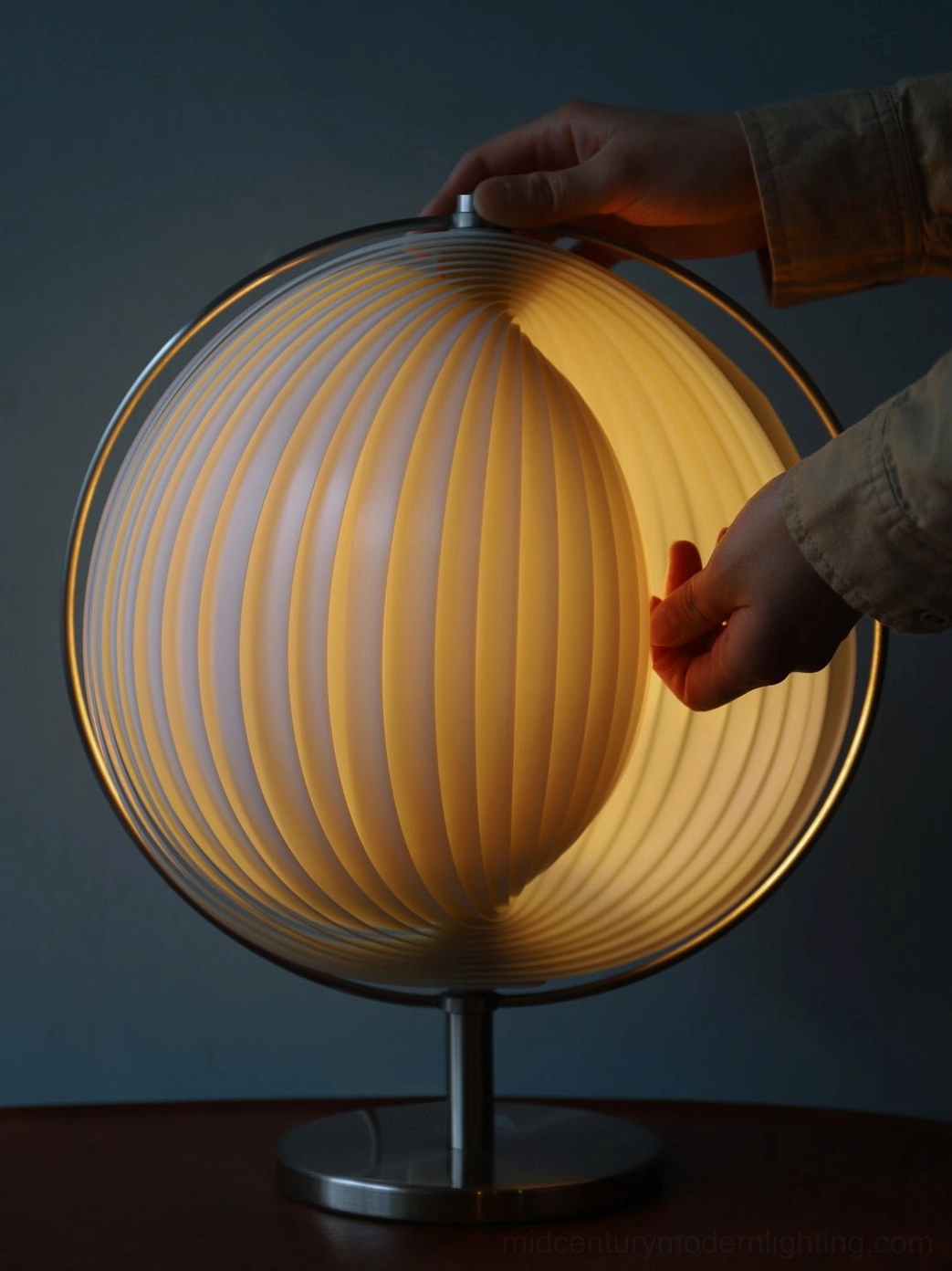 Moon Kare globe lamp shade with human hands adjusting rotatable mechanism, warm glowing light on dark tabletop
