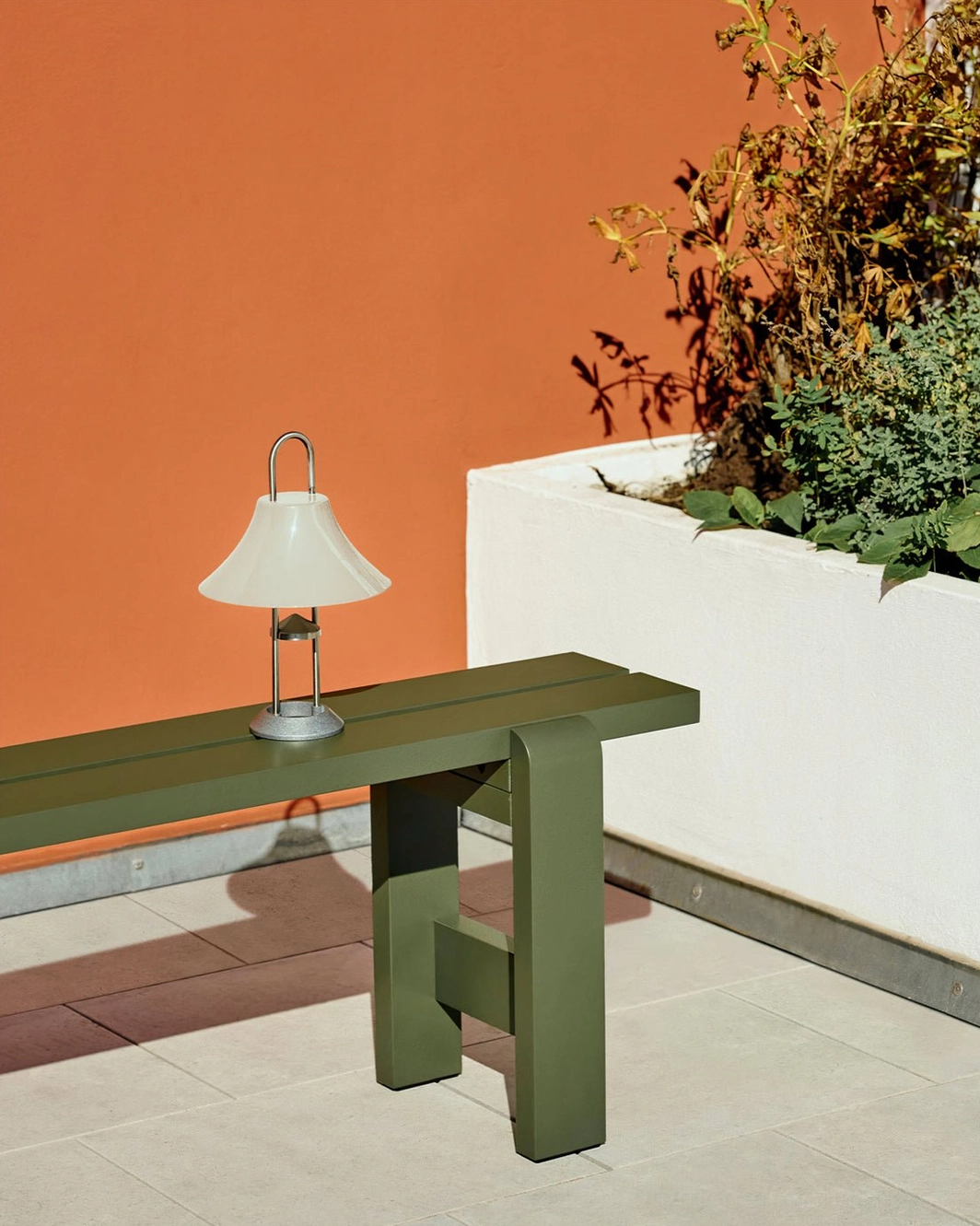 Portable battery-powered table lamp with carrying clip on outdoor bench against terracotta accent wall beside green furniture