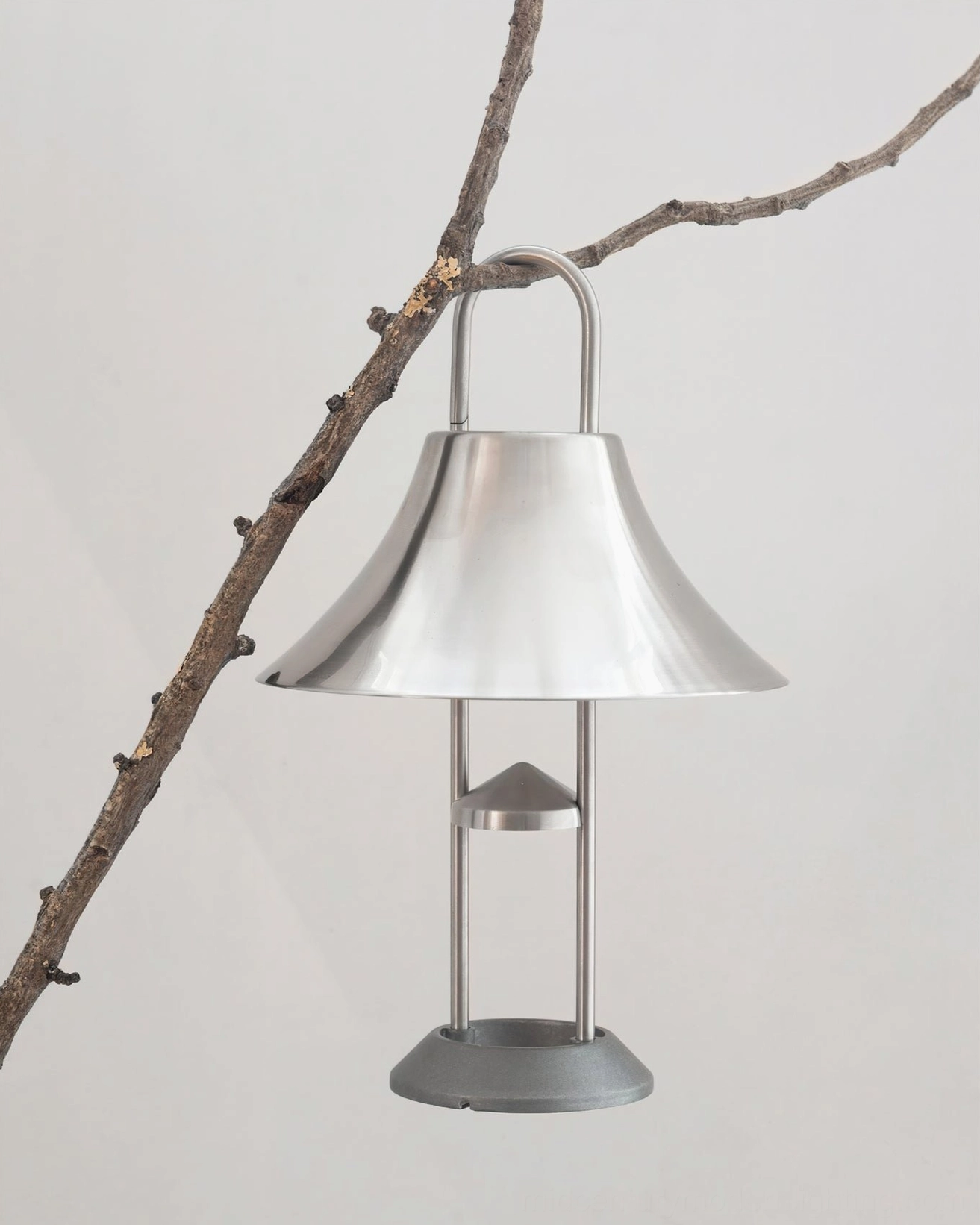 Brushed stainless steel Mousqueton portable table lamp with integrated clip handle outdoors against natural branch backdrop