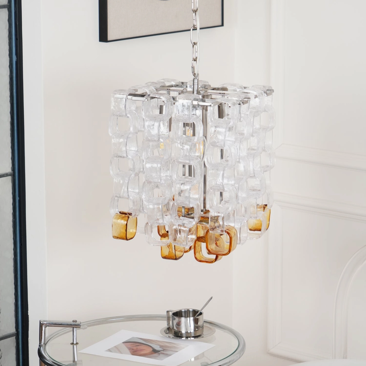 Side profile view of Elodie glass chandelier with chrome metal fixture, shown in minimalist white room with glass side table