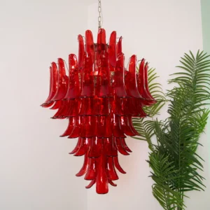 Red glass Murano chandelier installed in modern interior space with tropical plant, displaying translucent petal layers and warm lighting effect
