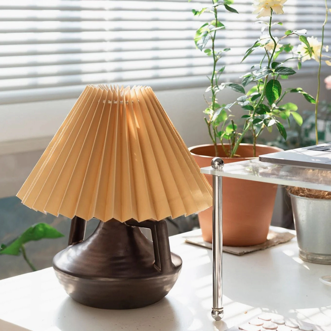 Navia table lamp on side table near window with blinds and fresh potted flowers