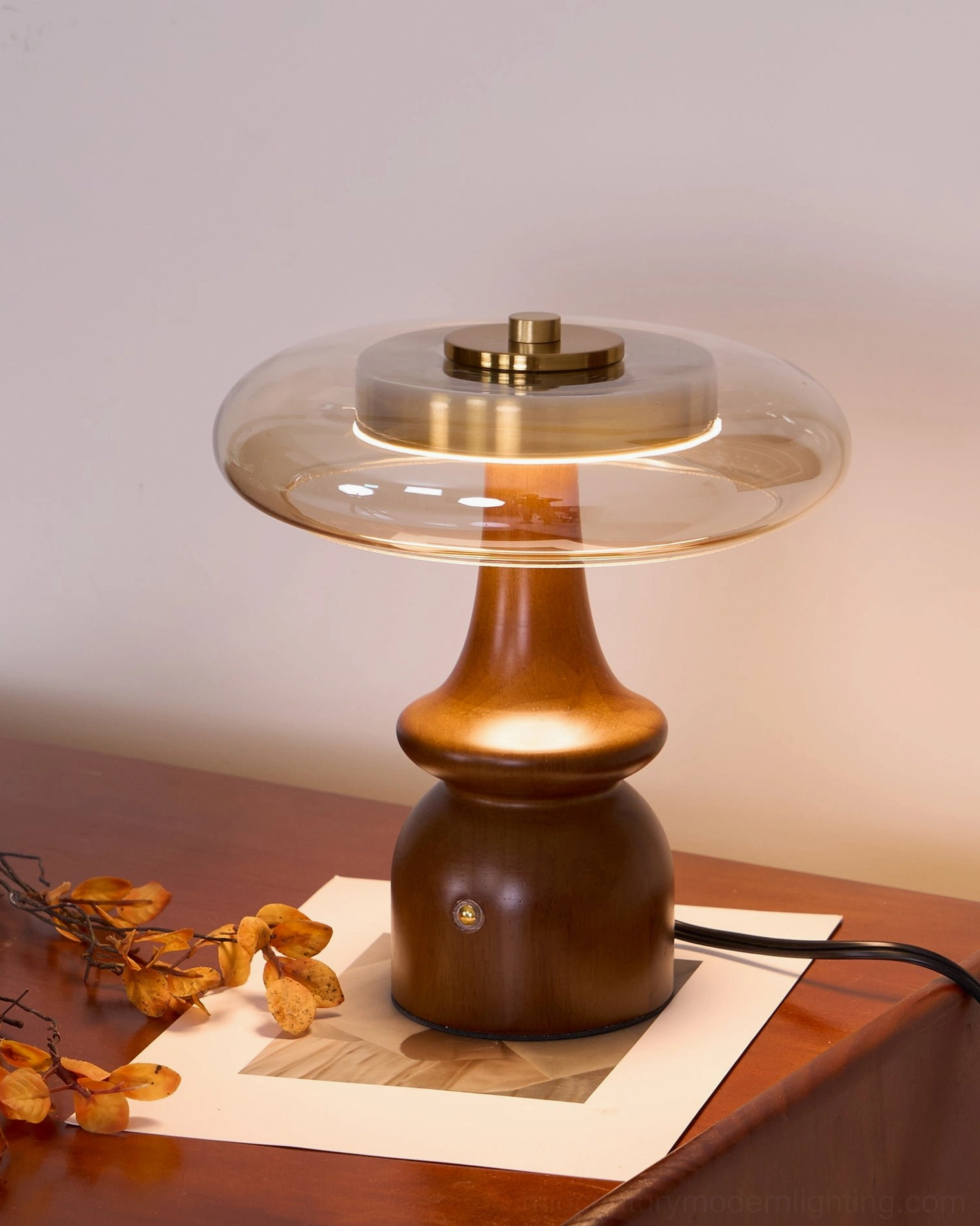 Nova table lamp lit from low left angle on wooden bedside table showing warm ambient light diffusion through glass shade