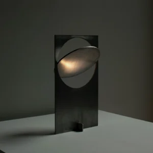 OBJ 01 brutalist table lamp three-quarter view with illuminated warm ambient glow, matte black minimalist metal construction, dark grey studio setting