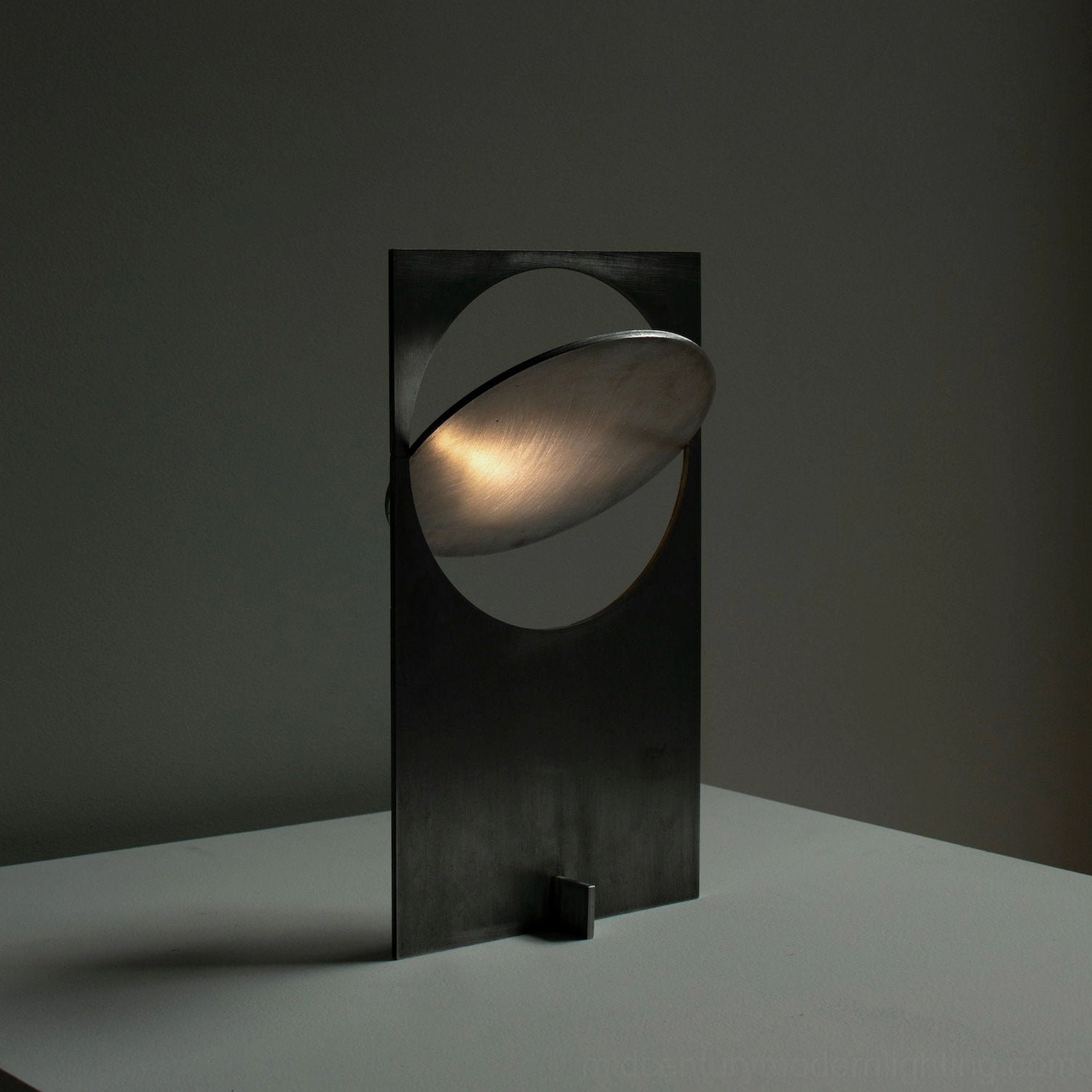OBJ 01 brutalist table lamp three-quarter view with illuminated warm ambient glow, matte black minimalist metal construction, dark grey studio setting