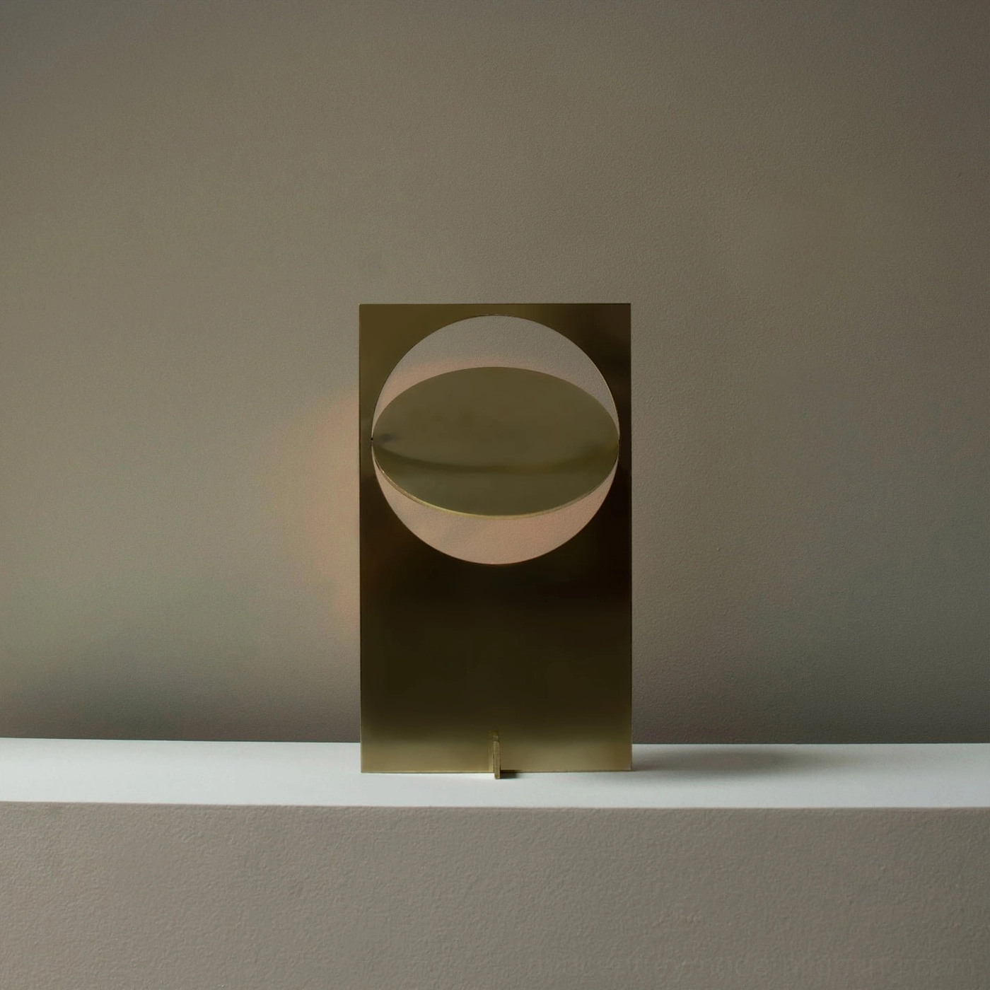 OBJ 01 table lamp brushed brass gold finish illuminated, three-quarter view with warm glowing light, wooden tabletop and neutral grey wall setting