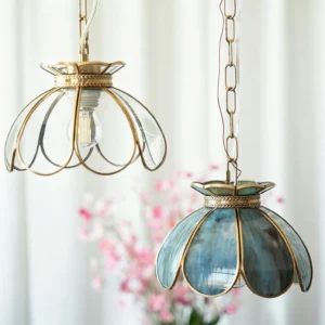 Two Odette pendant lights in blue-teal and clear glass variants displayed hanging together with white curtain and pink floral backdrop