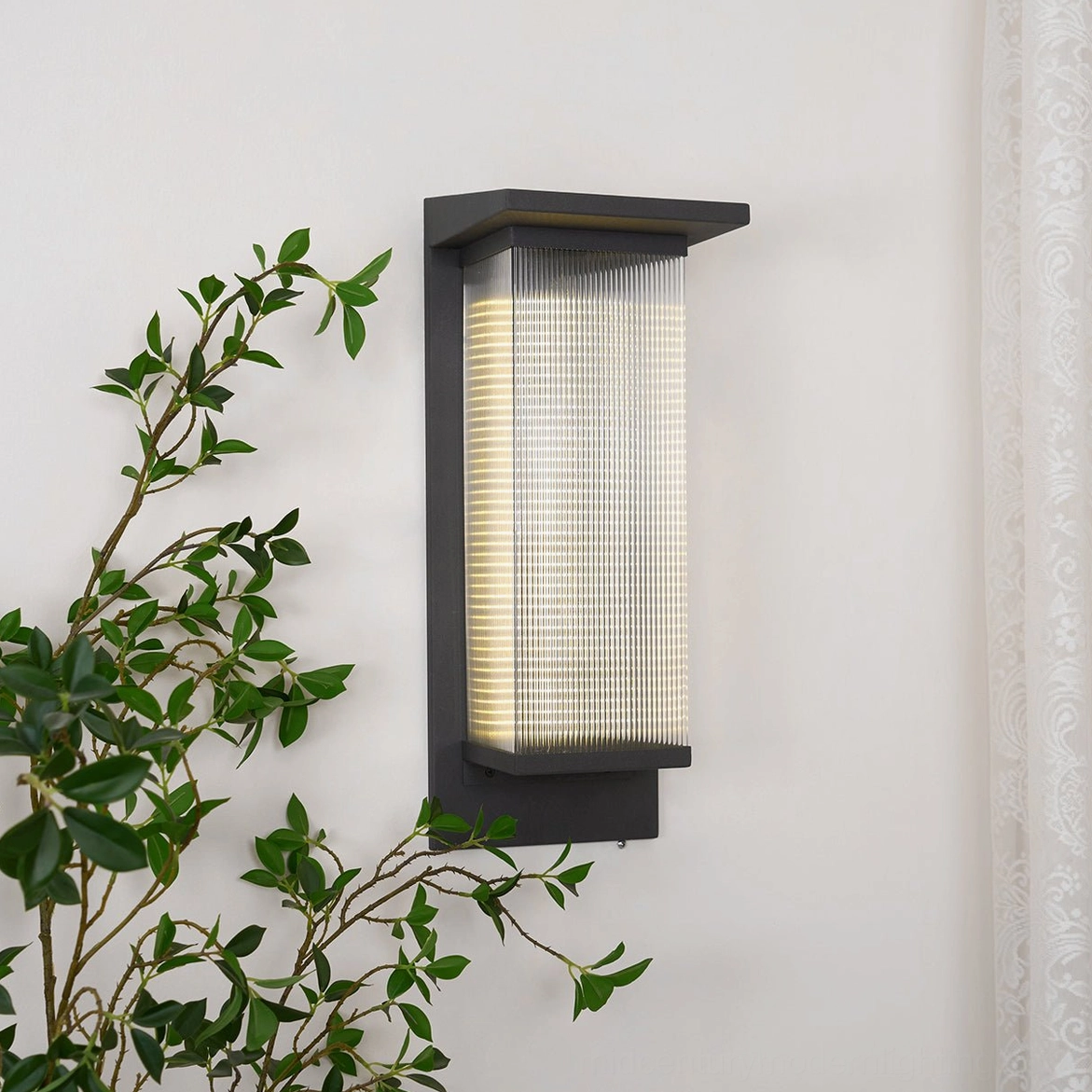 Oleron Box wall lamp side angle view illuminated, warm ambient light through textured glass, green foliage background, contemporary exterior lighting