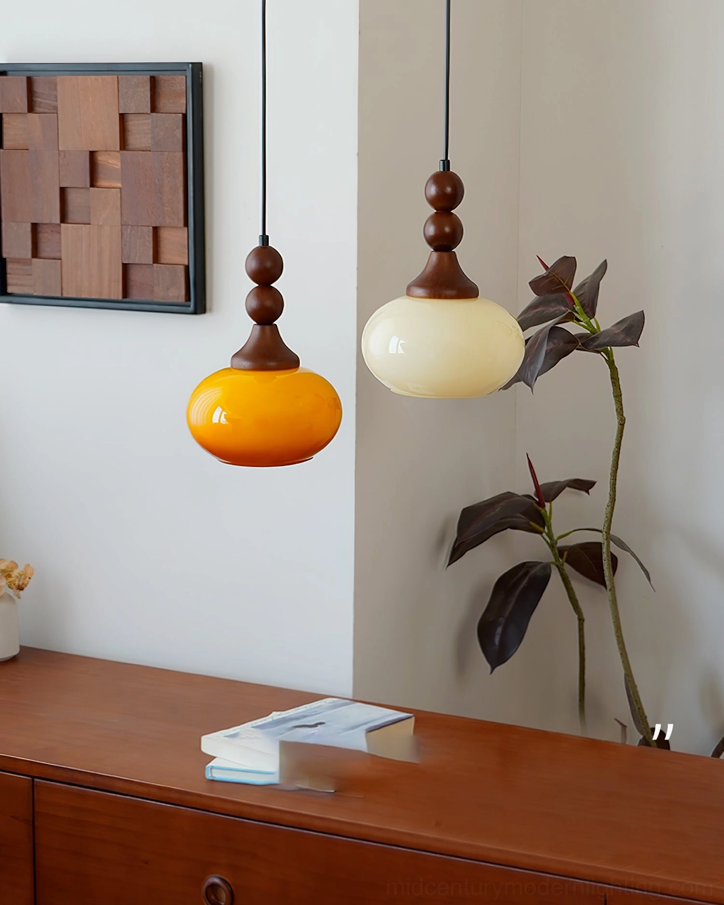 Two Orbique pendant lights in cream and amber glass displayed on wooden dresser with wall art and houseplant