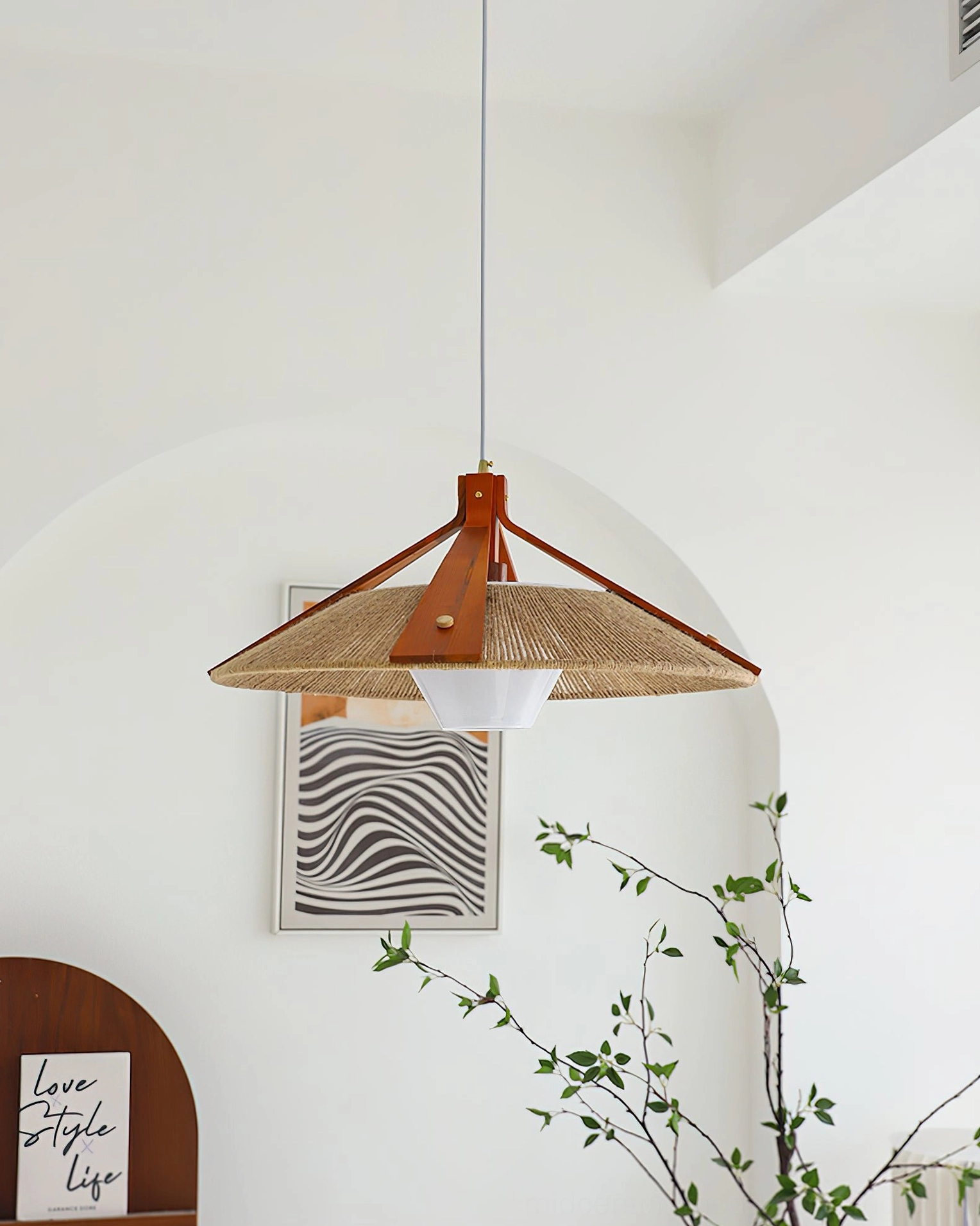 Artisanal woven pendant lamp hanging in indoor corner with green plants and wall decor, displaying warm ambient lighting and organic materials