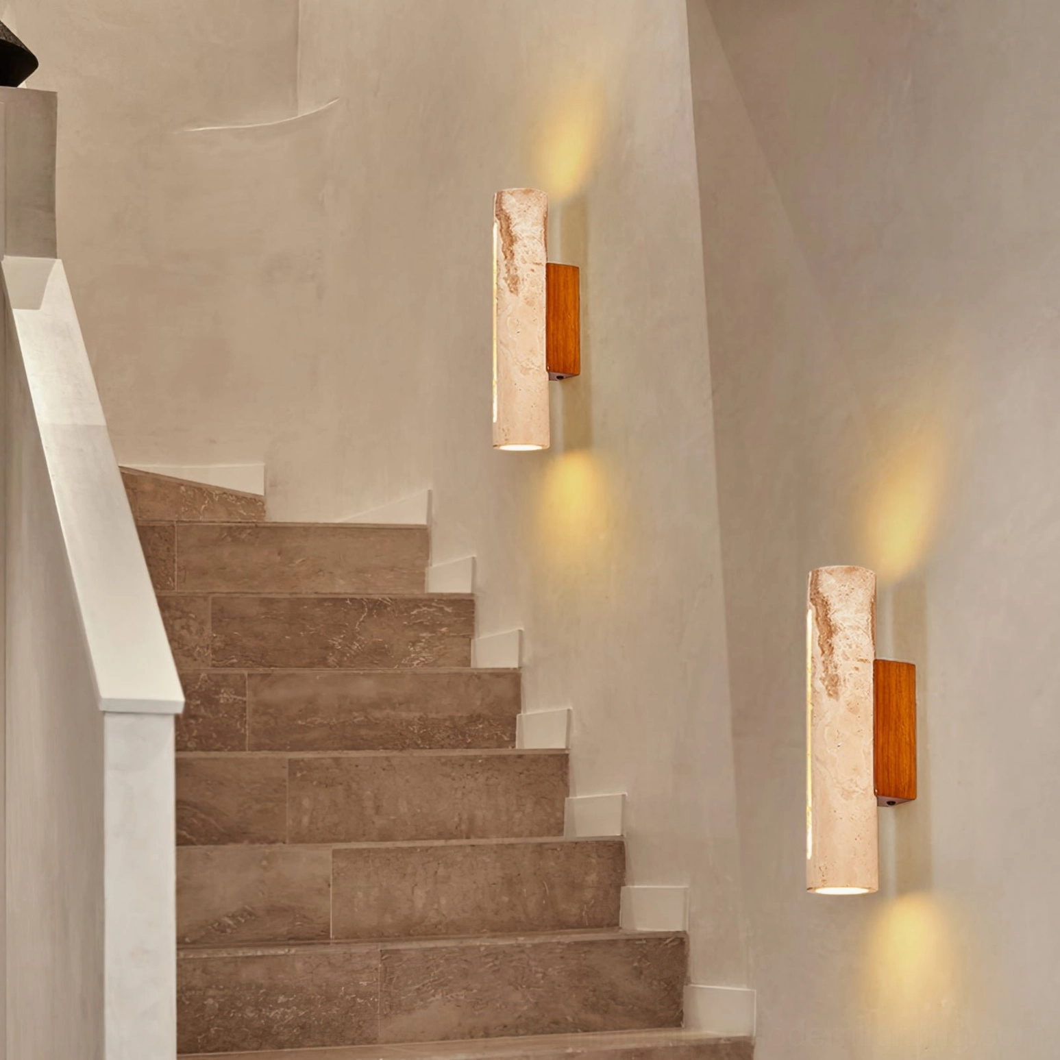 Pair of matching Lorelei travertine wall sconces on staircase with warm ambient glow and textured plaster walls