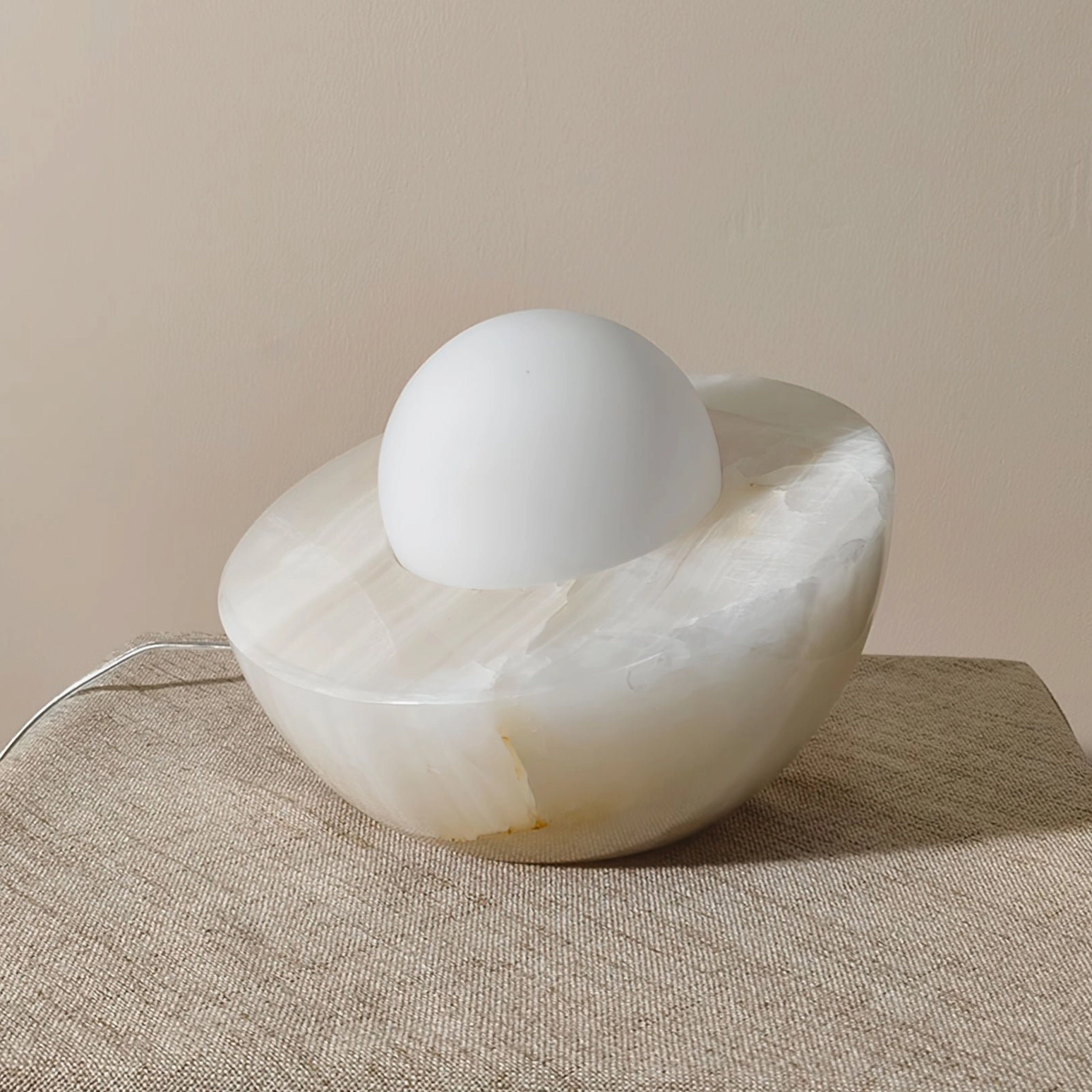 Piccola table lamp with natural onyx base and frosted sphere shade on linen fabric surface, warm ambient lighting, power cord visible