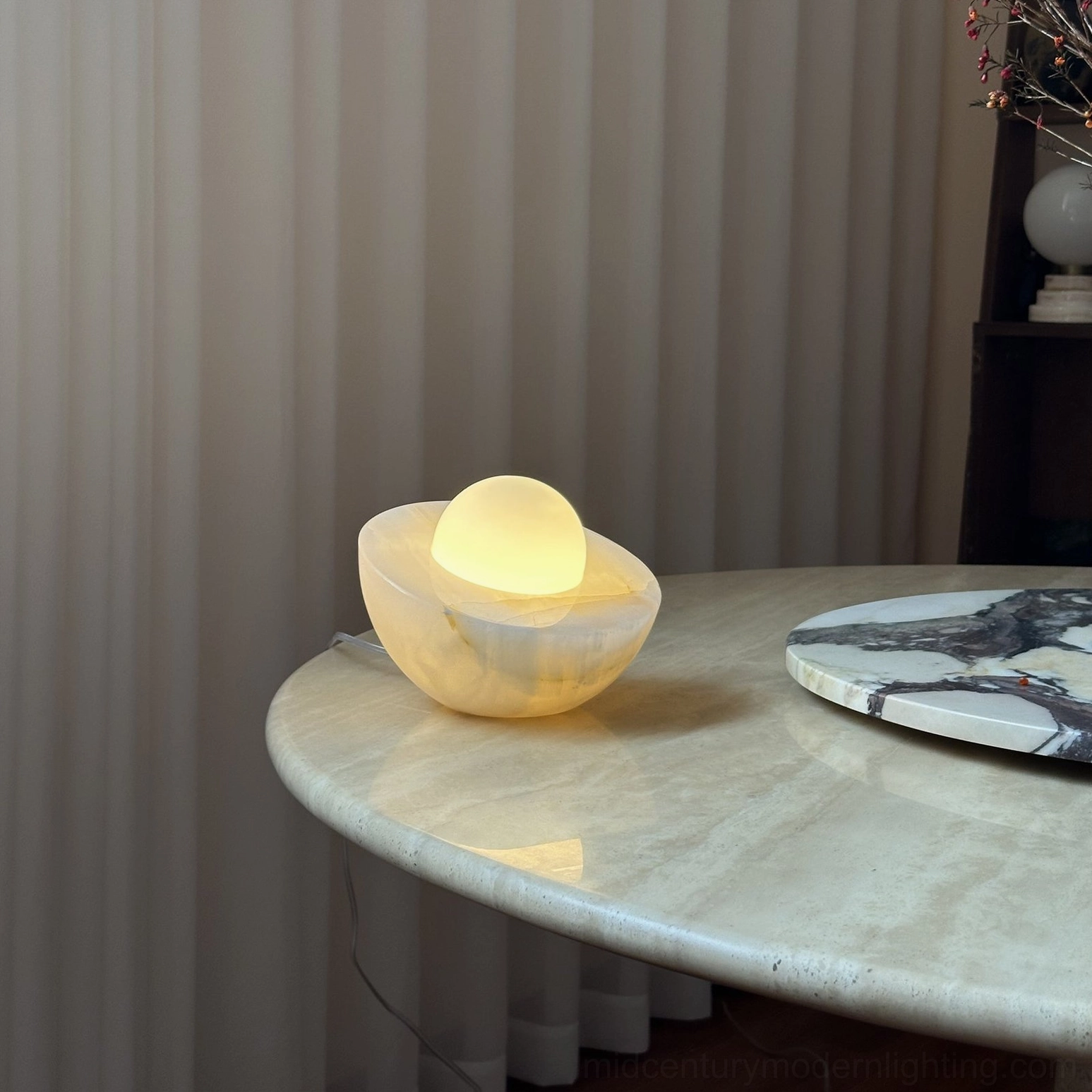 Piccola table lamp lit on marble surface with vertical curtain backdrop, warm light permeating through onyx base, sculptural form