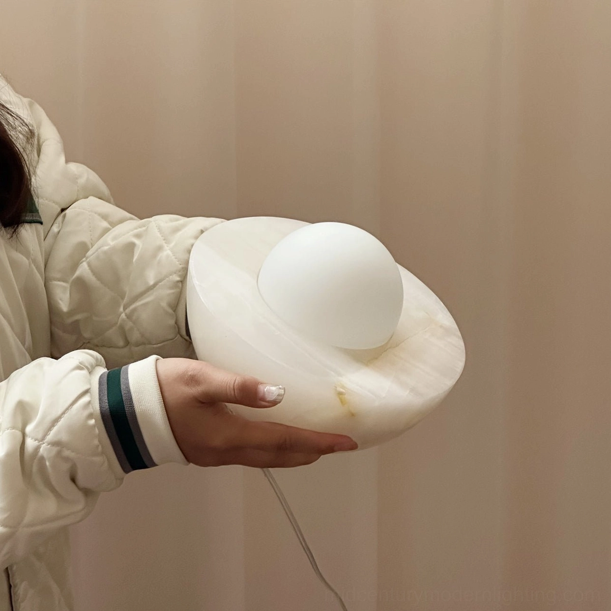 Woman holding Piccola table lamp to show scale and smooth marble surface, quilted jacket, neutral interior backdrop