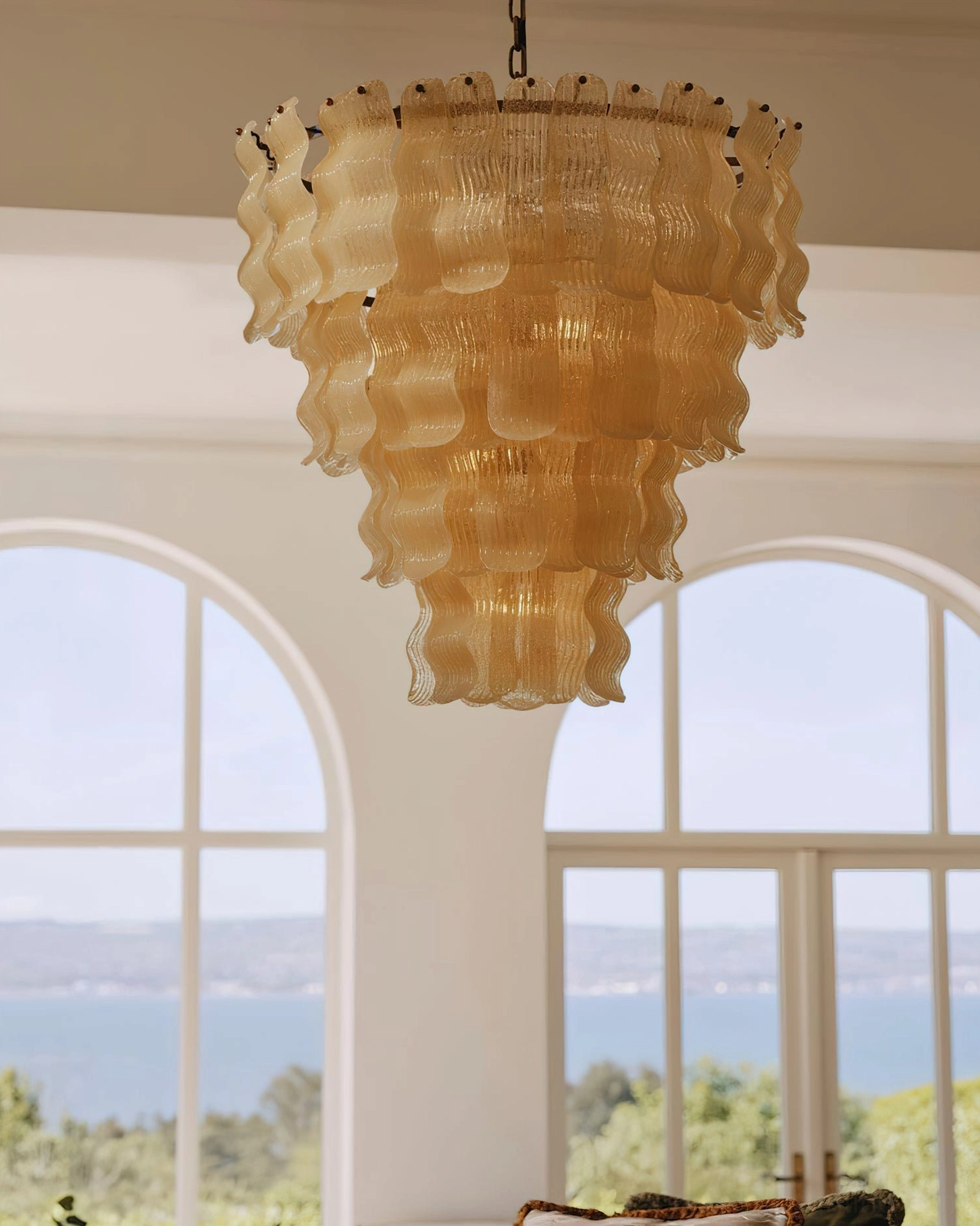 Illuminated Pollensa wave chandelier hanging in sunlit room with lake view, demonstrating light-diffusing graniglia effect