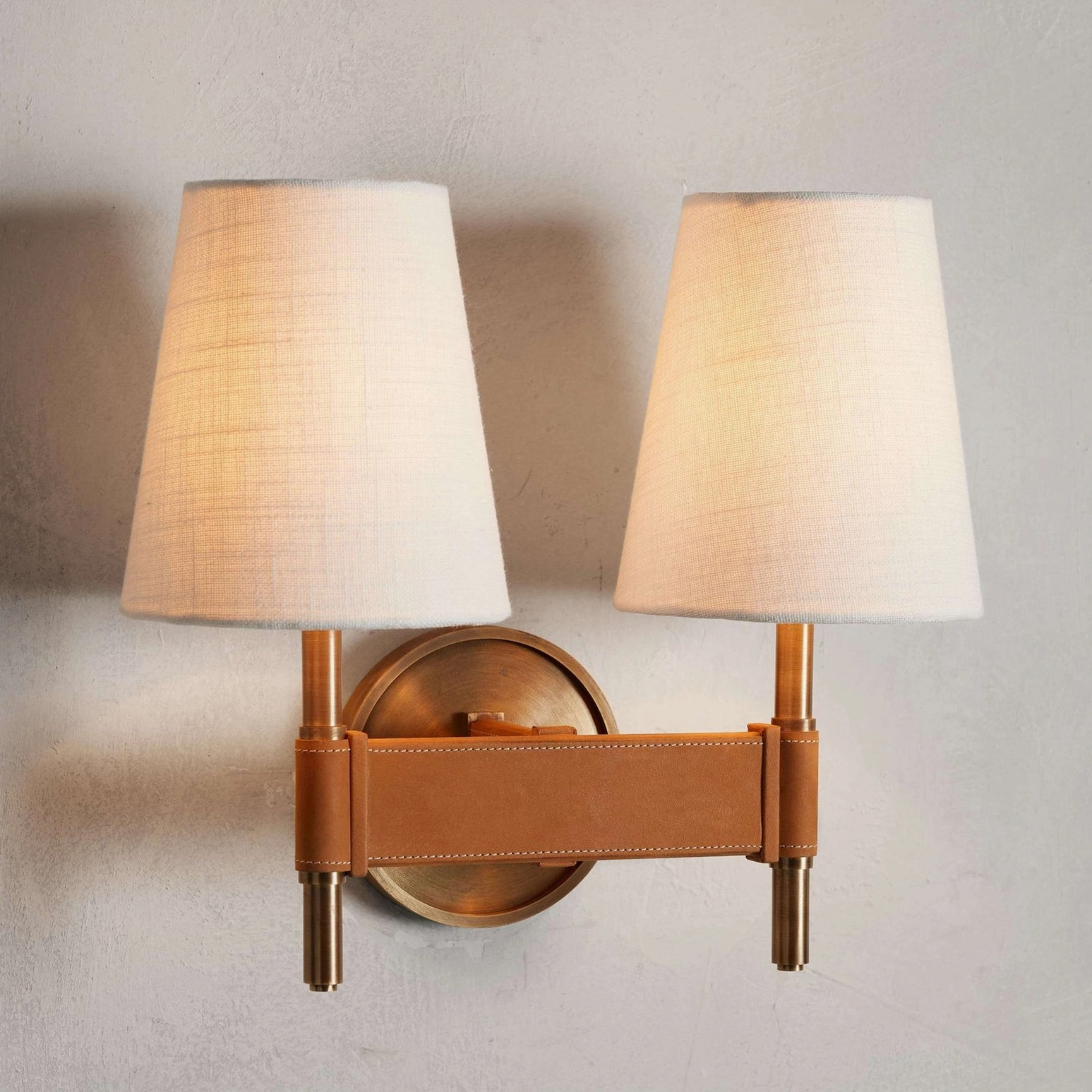 Prescot dual-head wall sconce mounted on home wall with both shades lit, displaying warm coordinated glow and leather-brass design integration