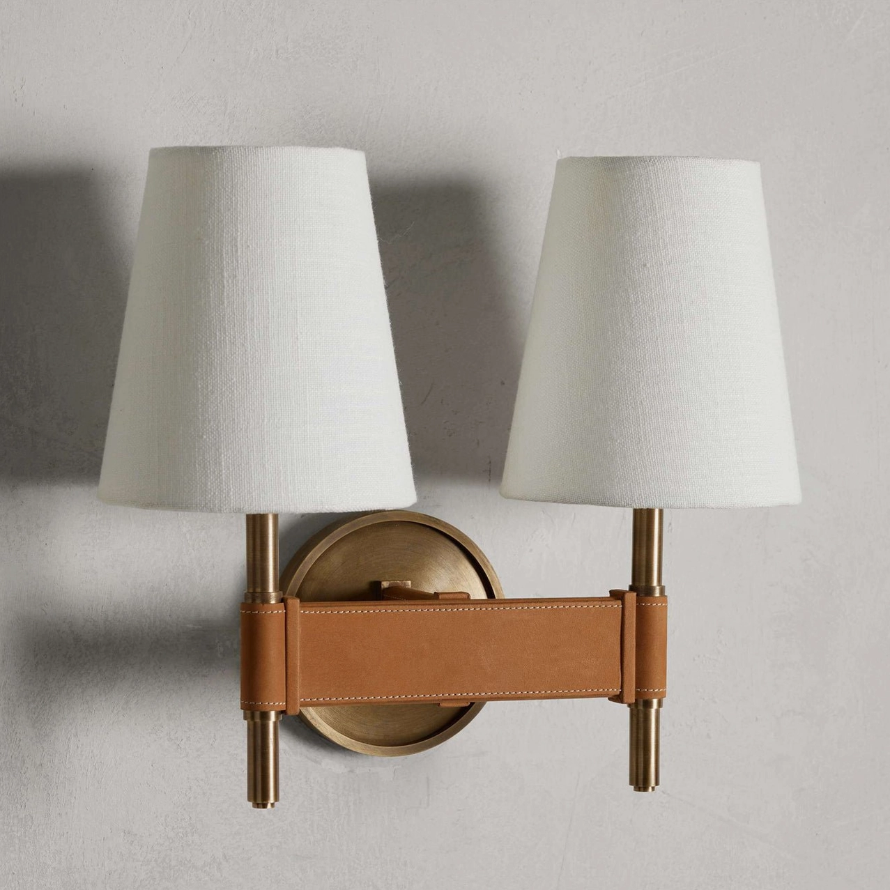 Dual-head Prescot wall sconce with two off-white fabric shades, matching brass hardware and coordinated tan leather crossbar on grey wall