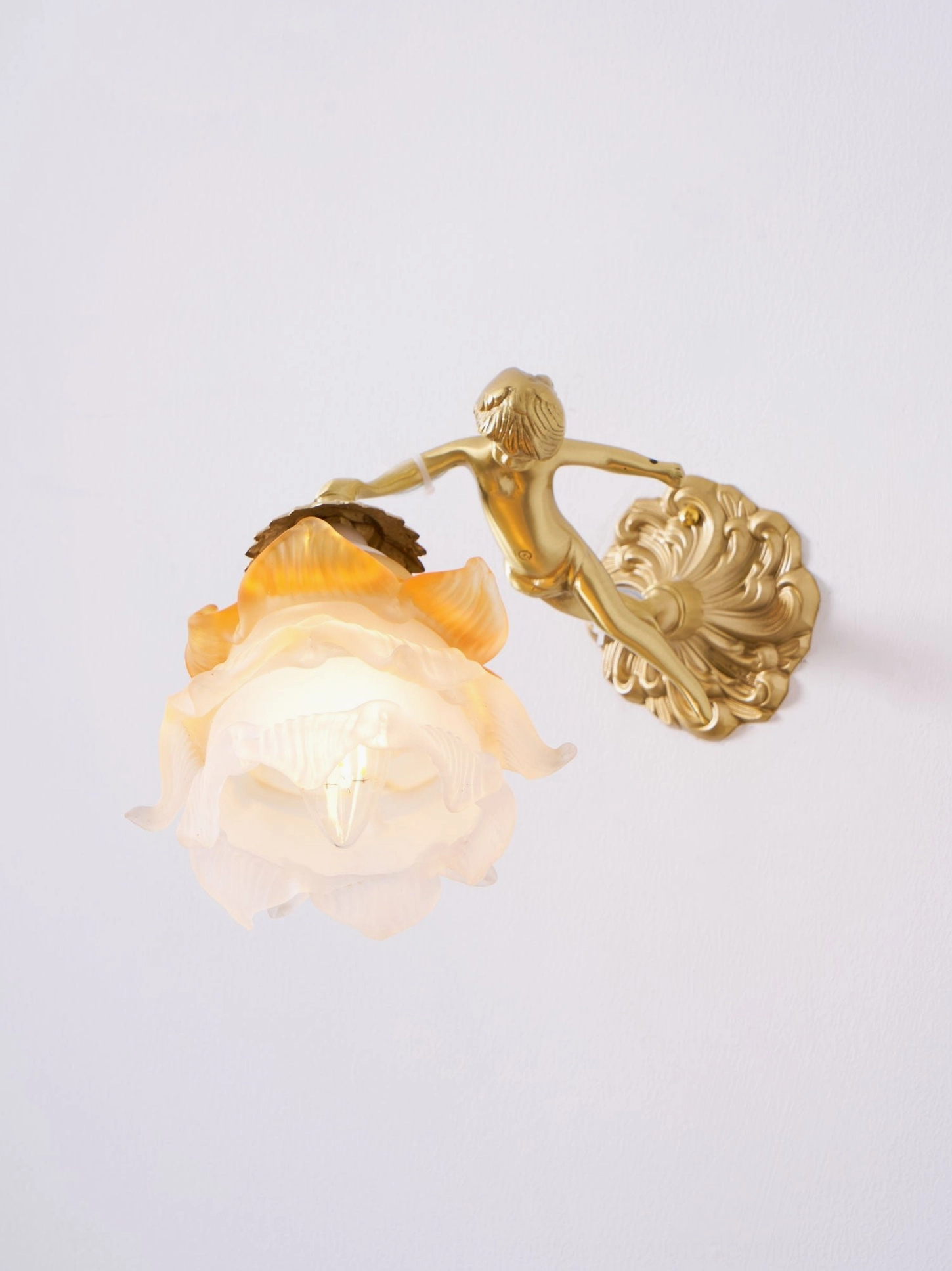 Illuminated putti cherub wall lamp beside home interior wall decor, frosted glass petal shade glowing warm, brass angel figure and romantic vintage styling