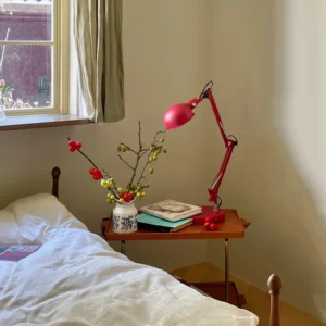 Red swing arm desk lamp positioned on wooden bedside table with floral arrangement and warm home decor