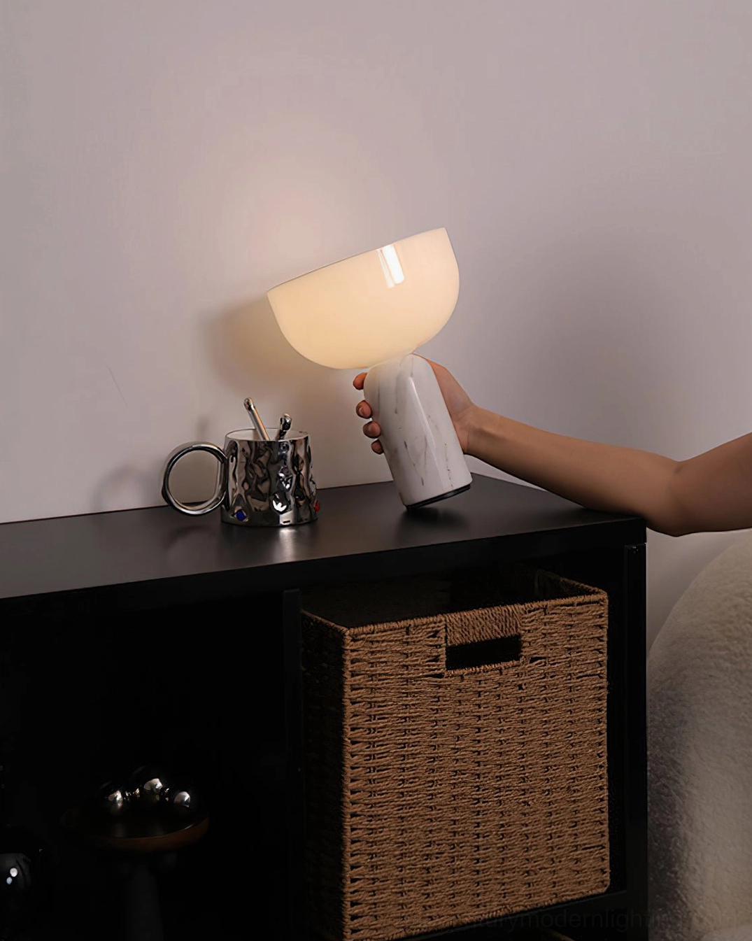 Hand holding white marble base Renard lamp with warm glow in bedroom shelf styling with metal cup