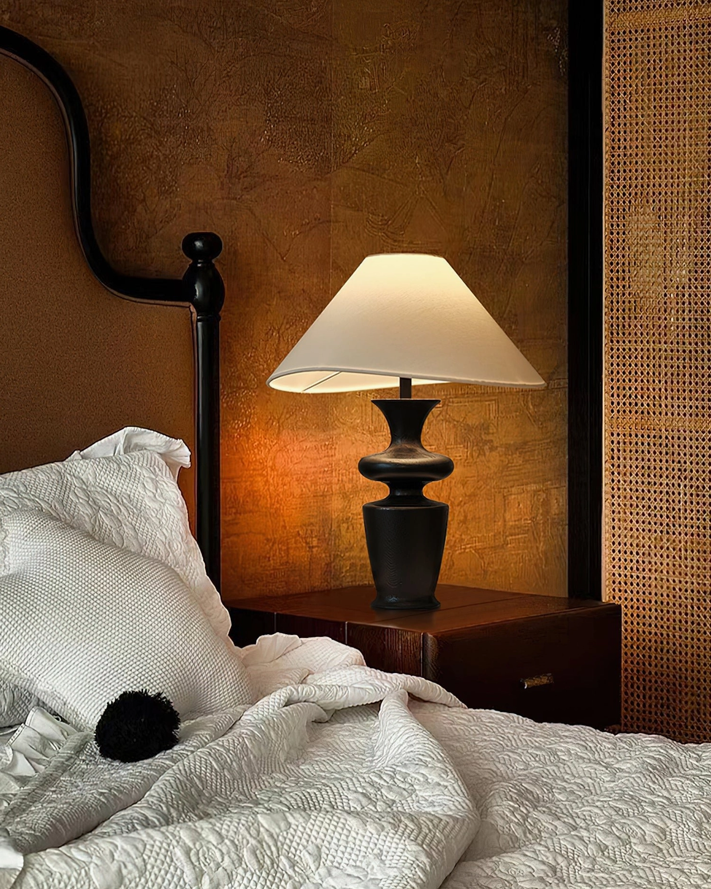 Low angle view of Rhodes table lamp by dark metal headboard with warm illumination over bedding