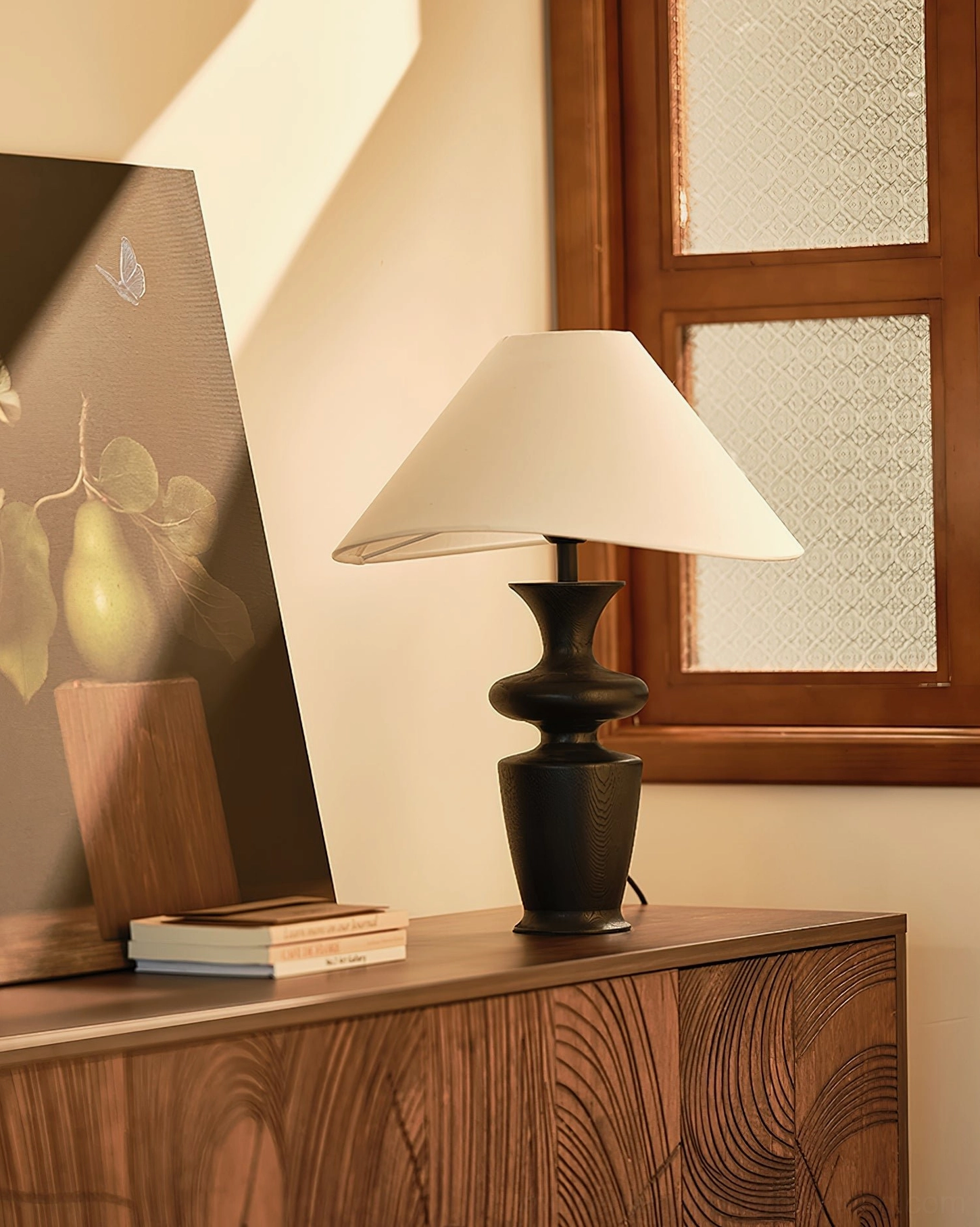 Side view of Rhodes sculptural table lamp with curved wooden base on carved dresser with stacked books