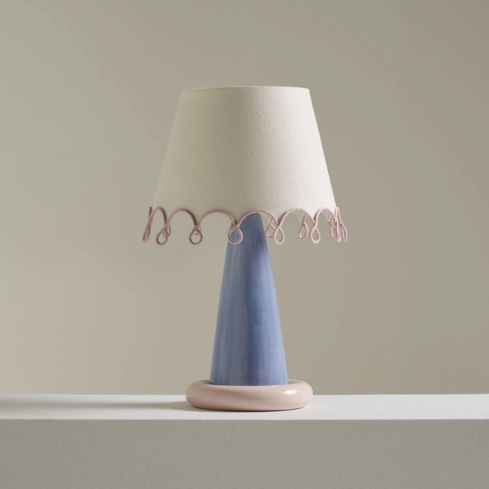 Close-up front view of modern ribbon table lamp showing blue ceramic body, pink ring base, and detailed pink rope scalloped trim on neutral tabletop