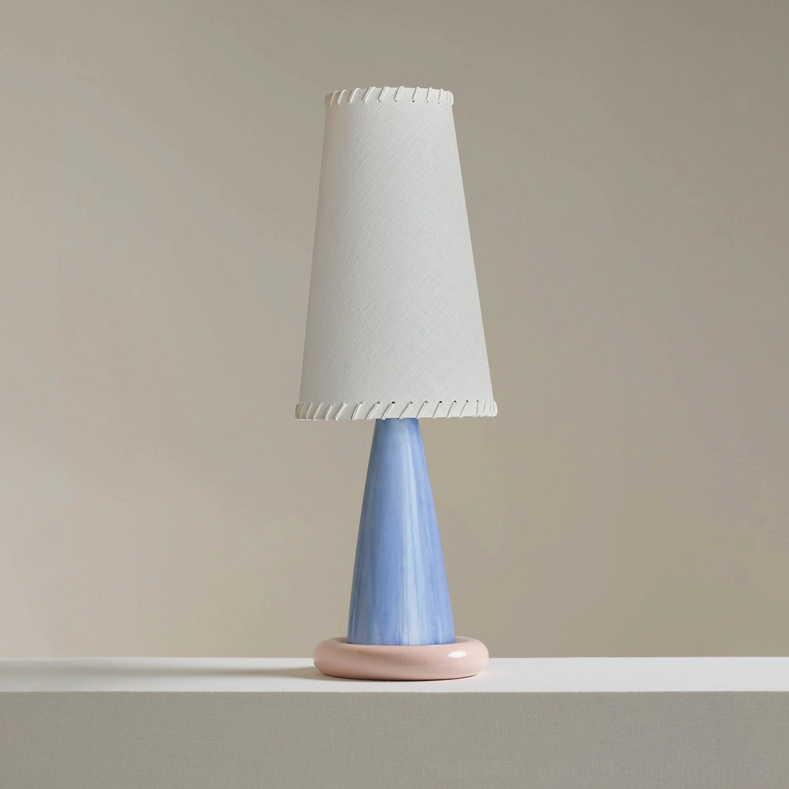 Full frontal shot of ribbon table lamp with tapered off-white fabric shade, light grey edges, and blue ceramic base with pink ring foot
