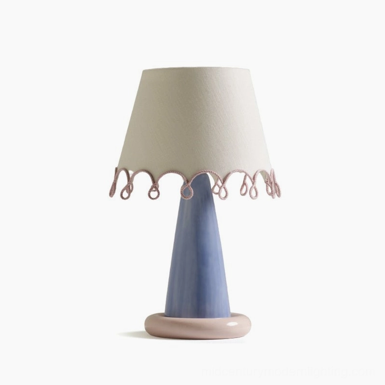 Flat lay product shot of ribbon table lamp with blue ceramic conical base, pink circular foot, and off-white shade with pink loop trim on white backdrop